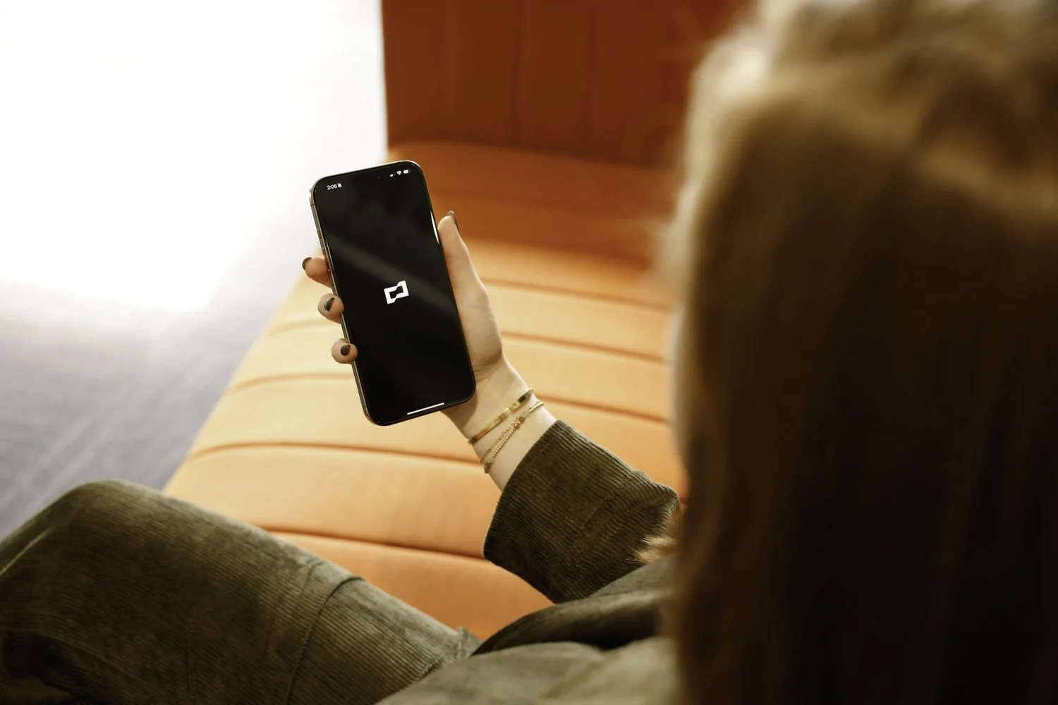 A woman with red hair holding a smartphone displaying an Brex logo, sitting on an orange sofa at Brex Headquarter in San Francisco, CA.