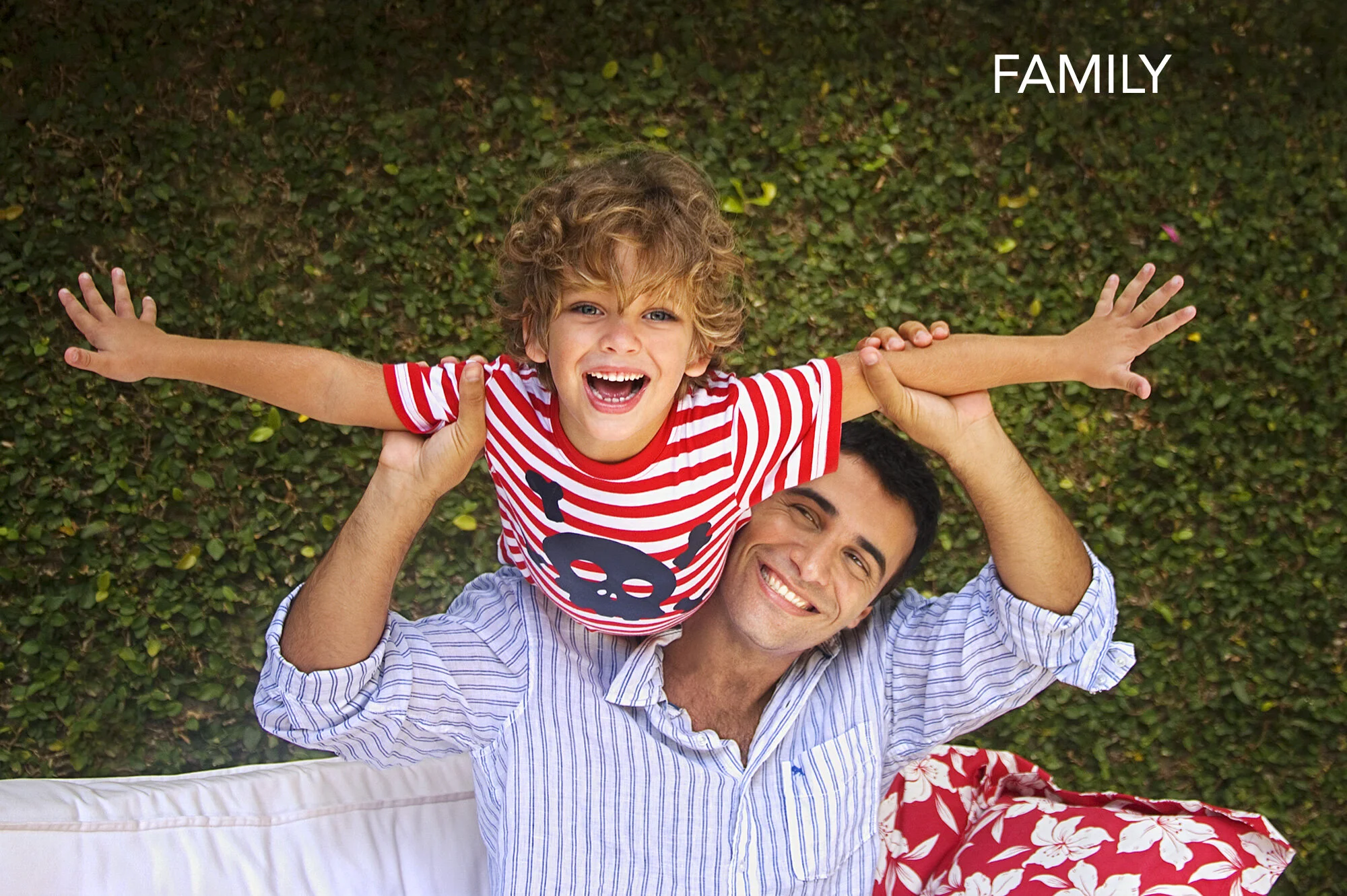 A joyful white boy with curly hair and his father, outside on green grass. The boy, wearing a red and white striped shirt with a skull design, is being held horizontally by the man, who is lying on the ground and smiling in Rio de Janeiro, Brazil.