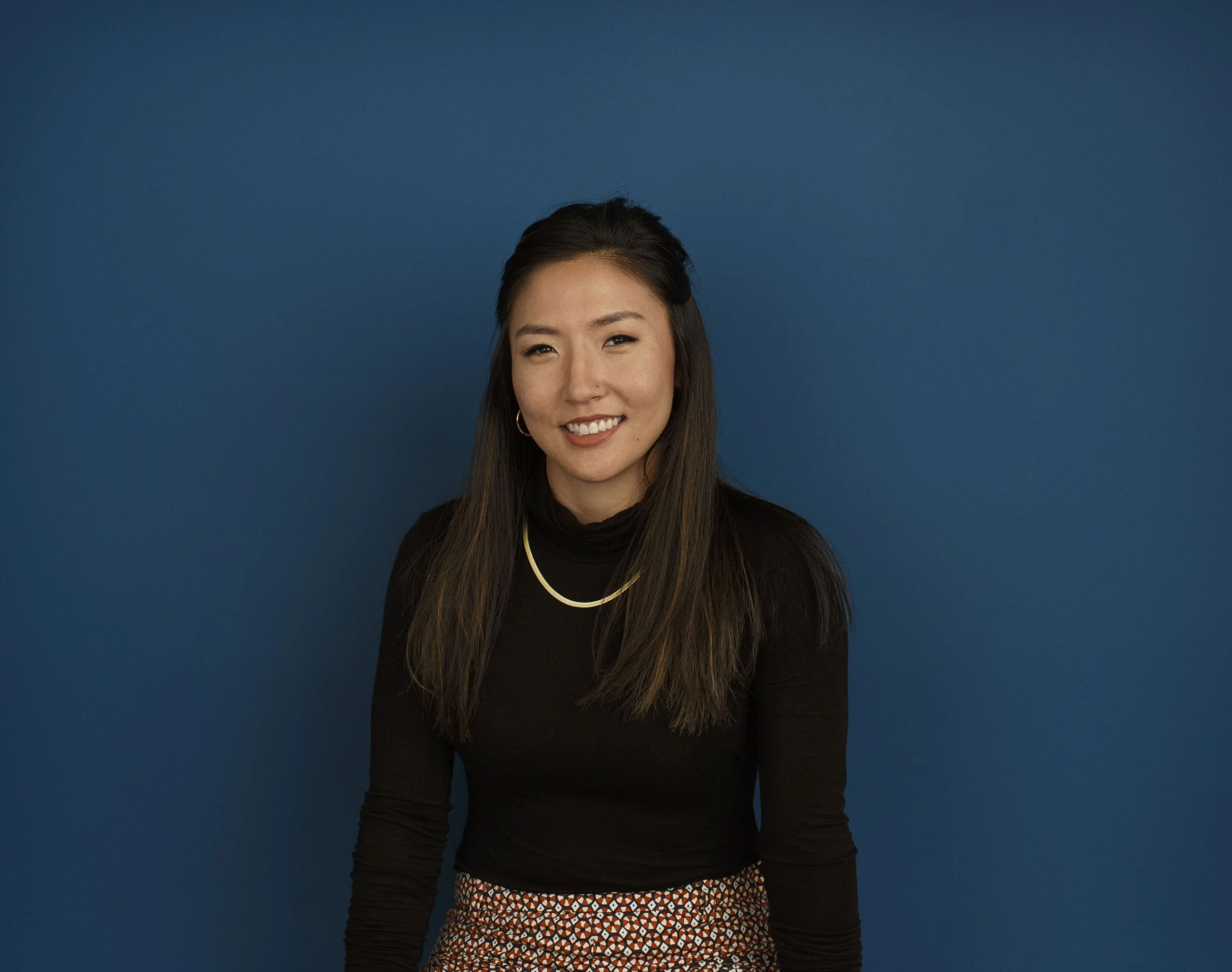 Portrait of a young Asian woman with long dark hair, smiling, wearing a black turtleneck, gold jewelry, and patterned pants, standing against a solid blue background in an architectural office in San Francisco, CA.