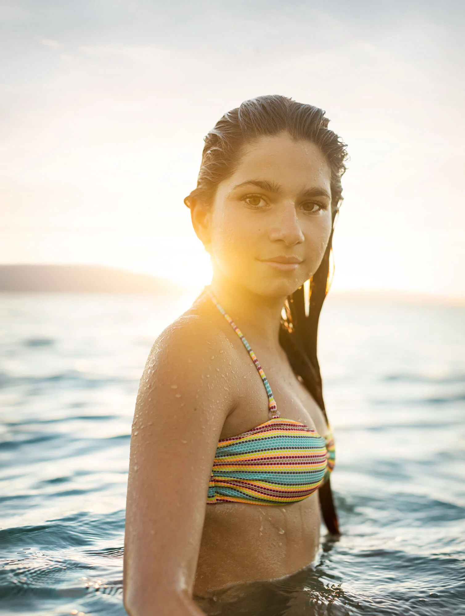 A white young woman with wet long hair in a striped bikini standing in the water during sunset with backlit light in Maui, Hawai'i.