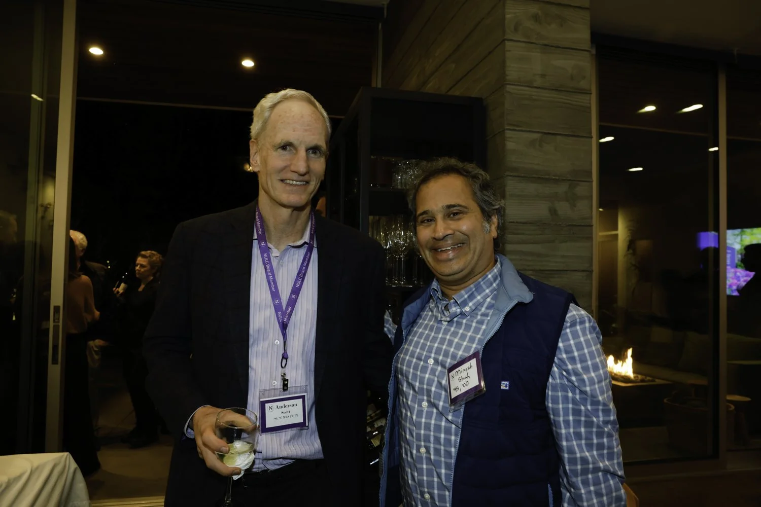 Two men at an indoor social event, one holding a glass of wine. The man on the left is tall, with light hair, wearing a checkered blazer with an event badge, and the man on the right has dark hair, wearing a blue vest and a checkered shirt. Both are 