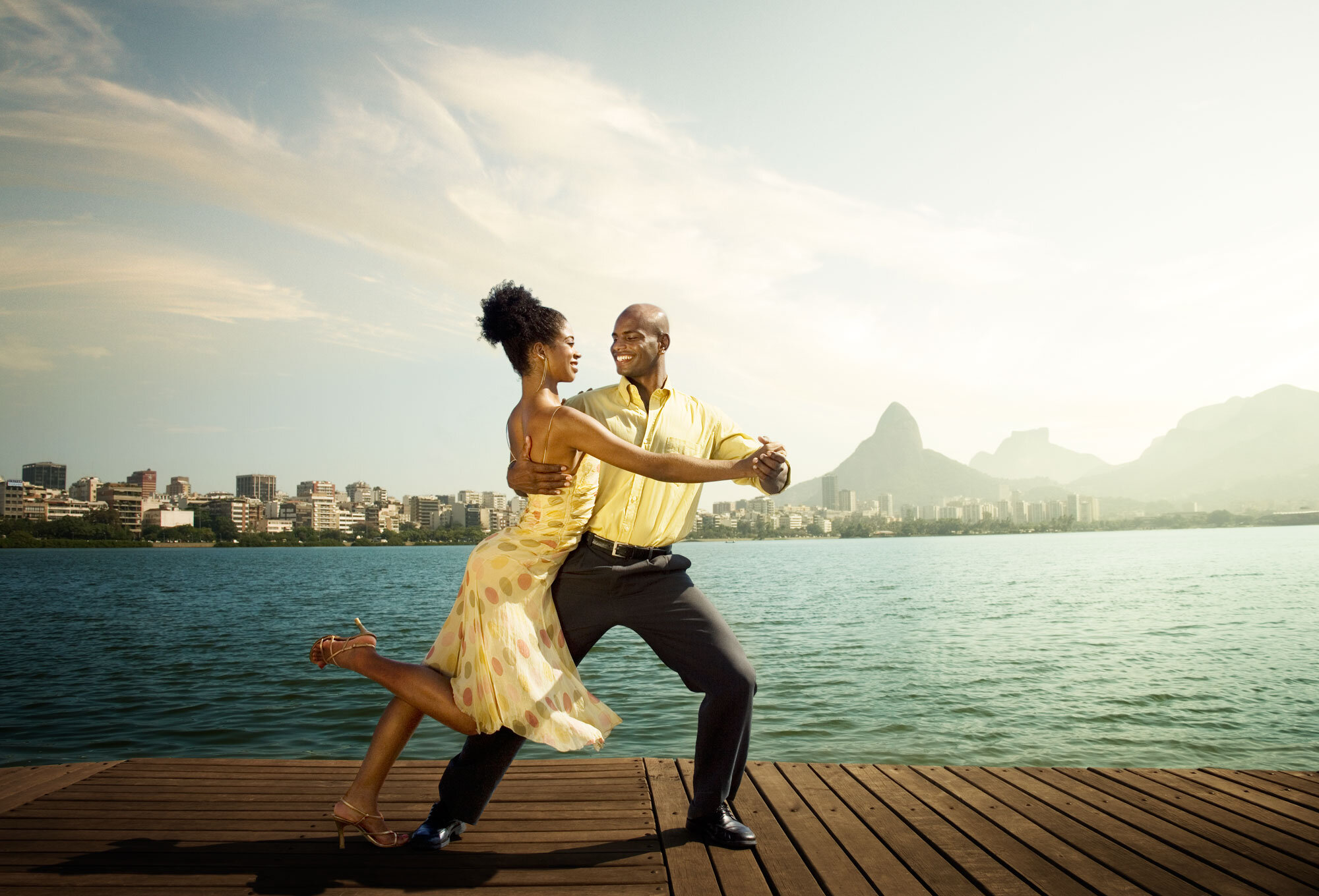 A Black Brazilian couple dancing on a wooden dock at Lagoa with a city skyline and mountains in the background, during sunset in Rio de Janeiro, Brazil.