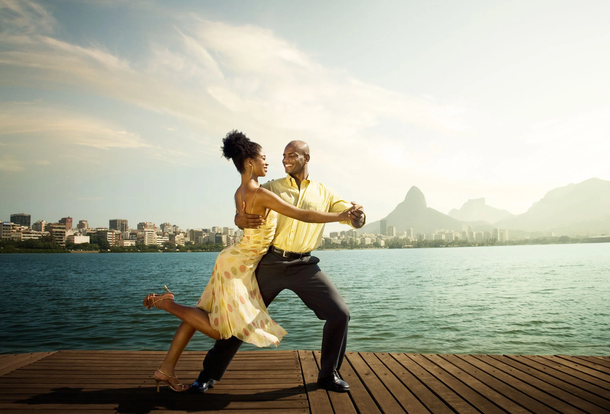 A Black Brazilian couple dancing on a wooden dock at Lagoa with a city skyline and mountains in the background, during sunset in Rio de Janeiro, Brazil.