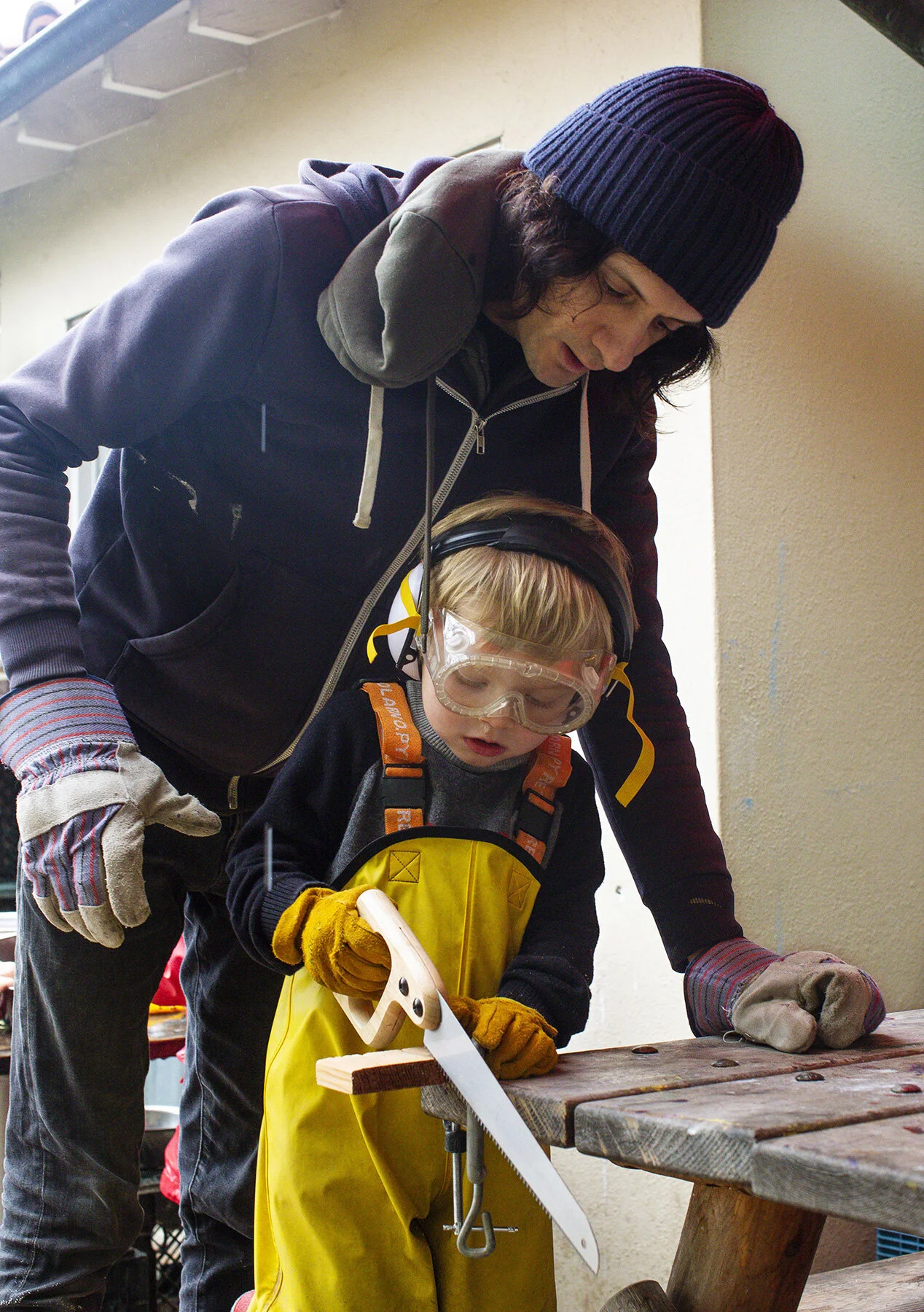 A teacher assists a young child wearing safety goggles and gloves in sawing a piece of wood outdoors. The man is leaning over, guiding the child's hand as he uses a hand saw at the The Scandinavian School in San Francisco, CA.