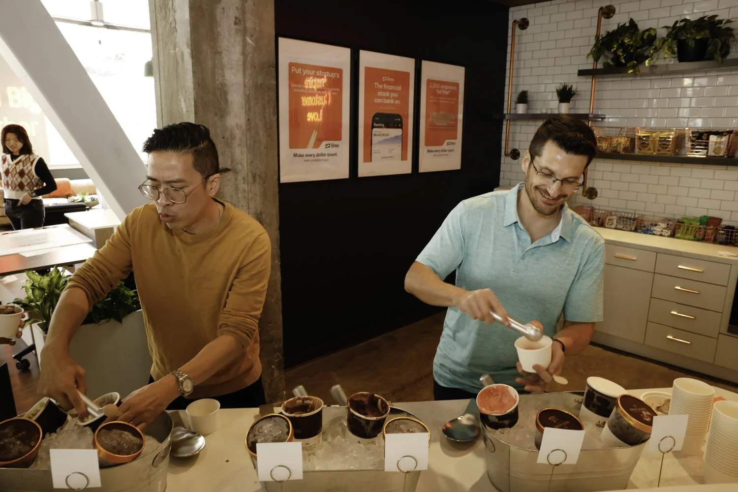 Two men serving ice cream in cups at a food event, with a woman in the background in a modern cafe setting Brex headquarter in San Francisco, CA.