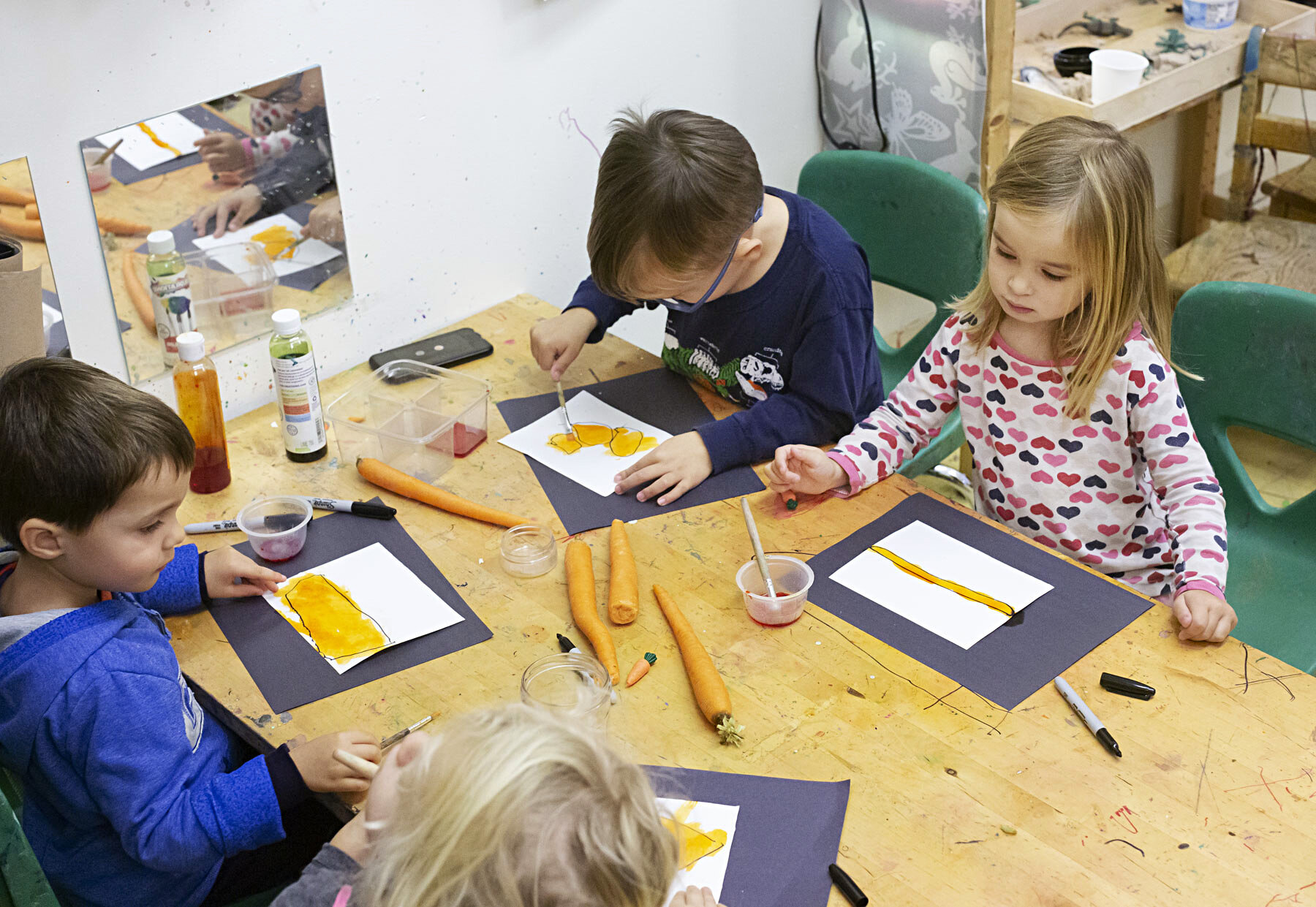 Children painting with carrots and yellow paint at a table in an art classroom at the The Scandinavian School in San Francisco, CA.