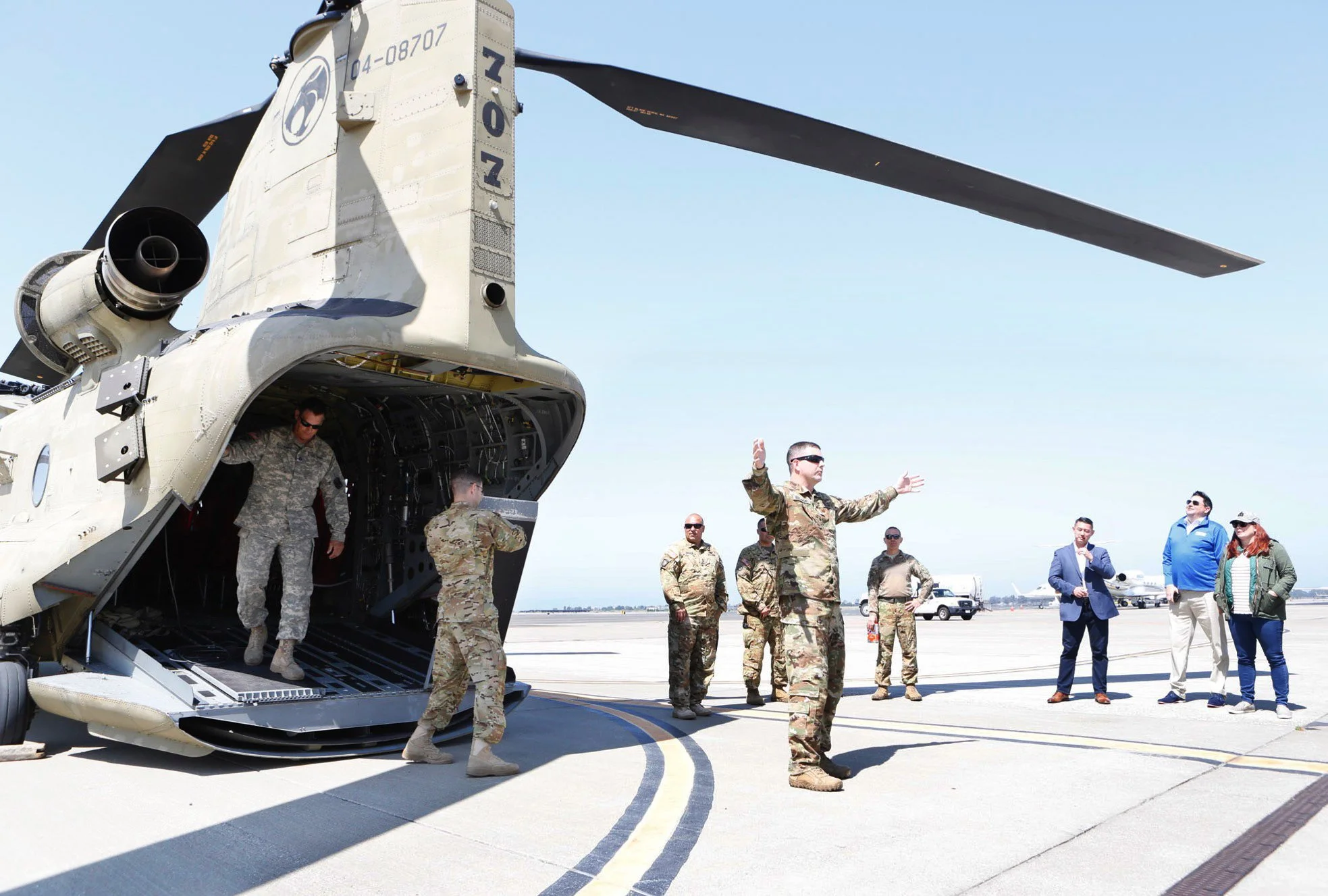 Military personnel with arms stretched out in front of a large military aircraft on an airfield during a clear day. A couple is standing on the sback of the aircraft in camouflage. Photographed  Salesforce Trailhead Military (Military Veteran Recruit