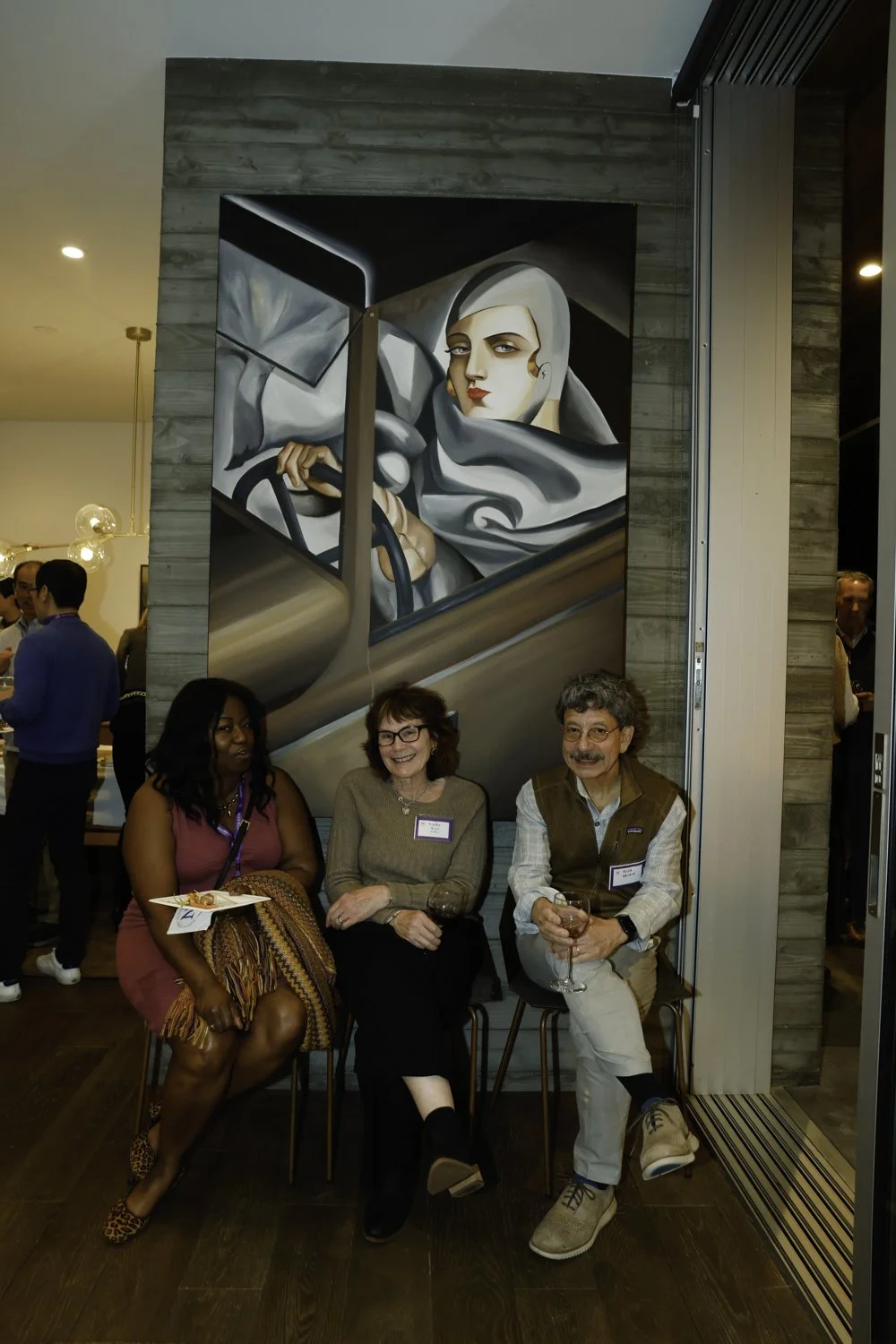 Three people sitting in front of a large painting of a woman in a car. The woman in the painting has blonde hair and is looking at the viewer. The setting appears to be a social event or gathering at Northwestern fundraiser in San Francisco, CA.