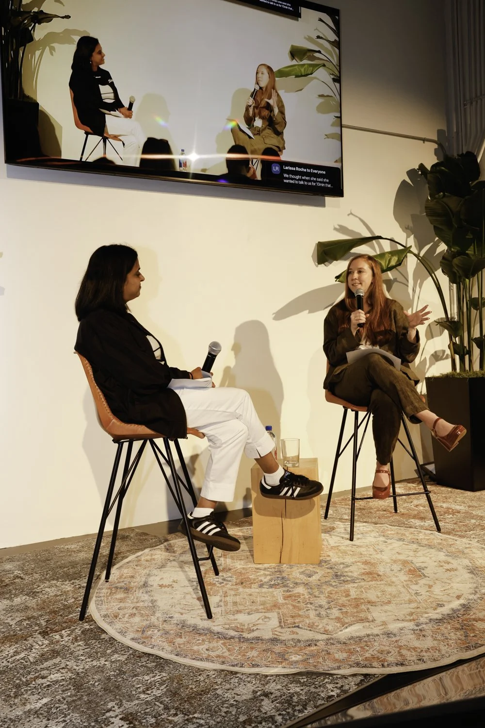Two women seated on stage in conversation, one with a microphone, a large display screen showing their live video feed, and a potted plant nearby, in an indoor event setting Brex headquarter in San Francisco, CA.