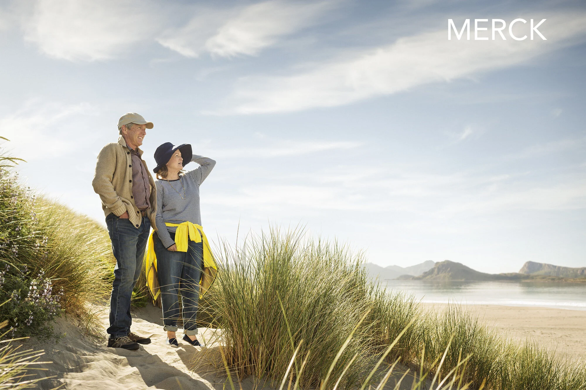 Two elderly people, a man and a woman, standing on a sandy beach among tall grasses, looking out at the ocean and distant mountains under a partly cloudy sky. The woman is wearing a wide-brim hat, striped shirt, and has a yellow jacket tied around he