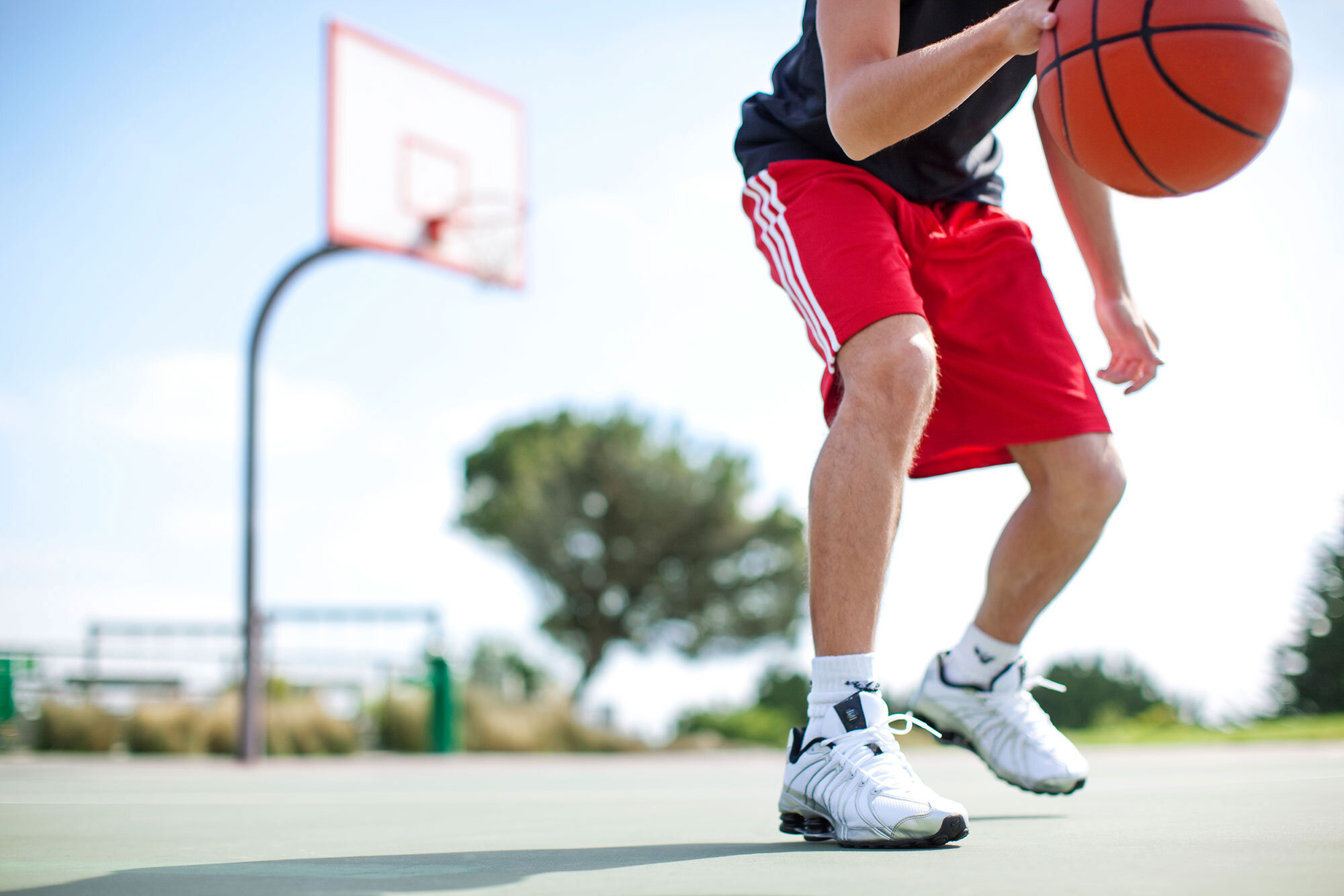 A young Latino man playing basketball on an outdoor court, dribbling the ball while wearing a black shirt, red shorts, and white athletic shoes, with a basketball hoop in the background in San Francisco, CA