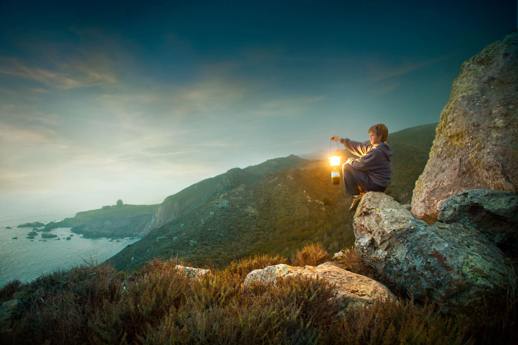A young boy sitting on rocks at sunset, holding a lantern glowing, overlooking a coastal landscape with hills and the ocean in Marin, CA.