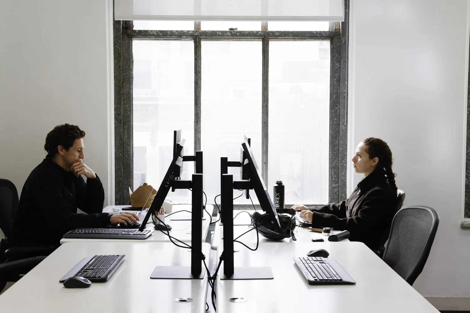 Two people working at separate desks with computers in an office, positioned back-to-back, facing each other, with a large window in the background at Brex Headquarter in San Francisco, CA.