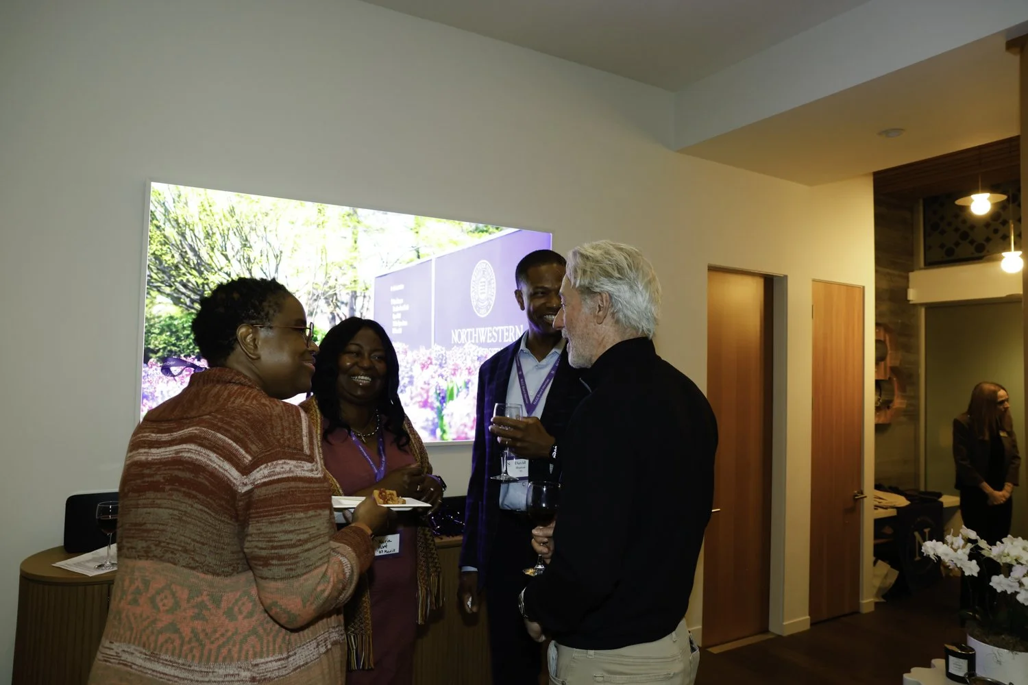 Four people engaging in conversation at a social event in a room with wooden doors, a large digital screen, and warm lighting. One woman is holding a plate of food, and two men are holding wine glasses at Northwestern fundraiser in San Francisco, CA.