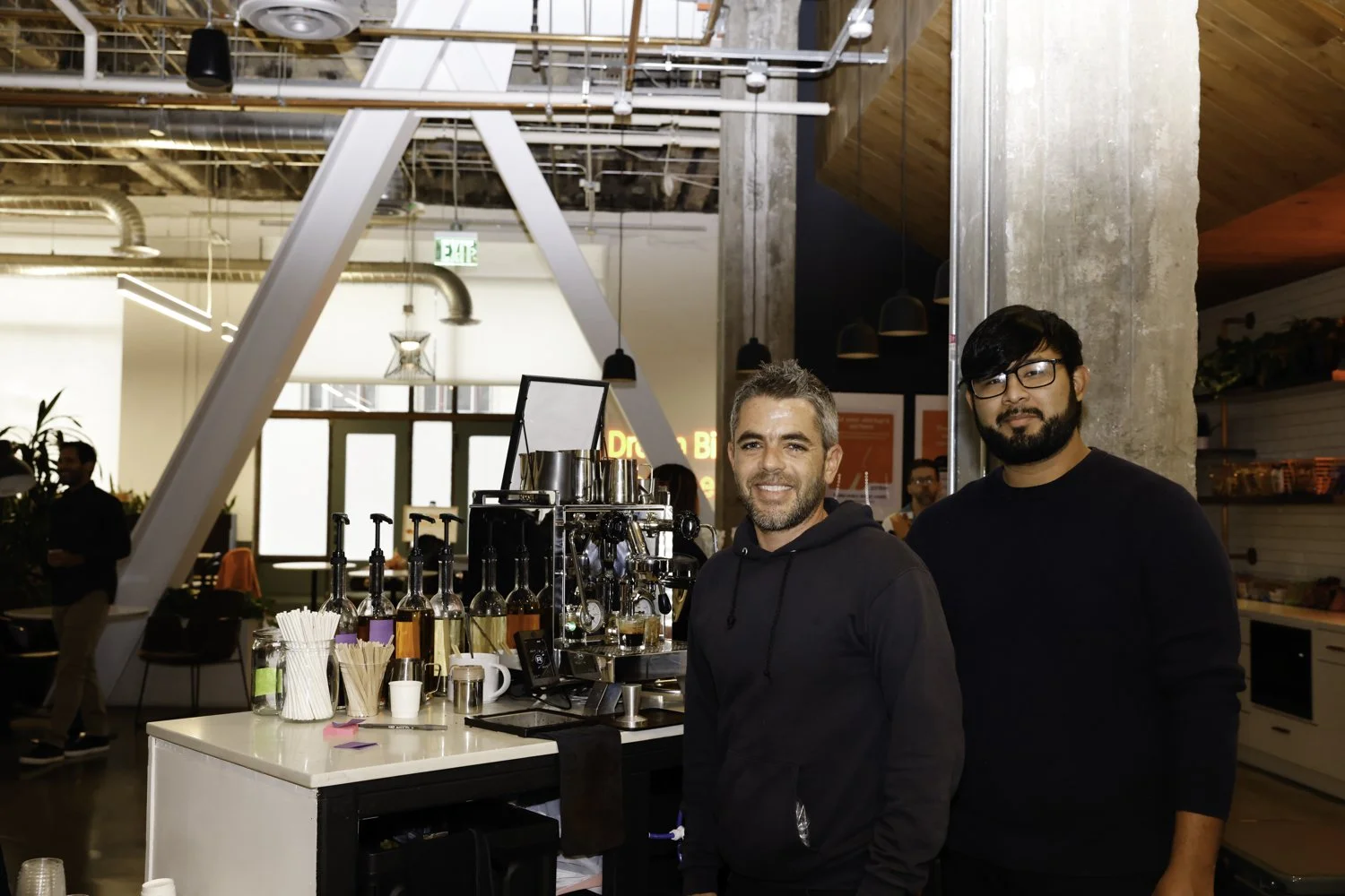 Two men standing behind a coffee bar in a modern cafe. One has a beard and gray hair, the other is wearing glasses and has a beard. Coffee equipment and bottles are on the counter Brex headquarter in San Francisco, CA.