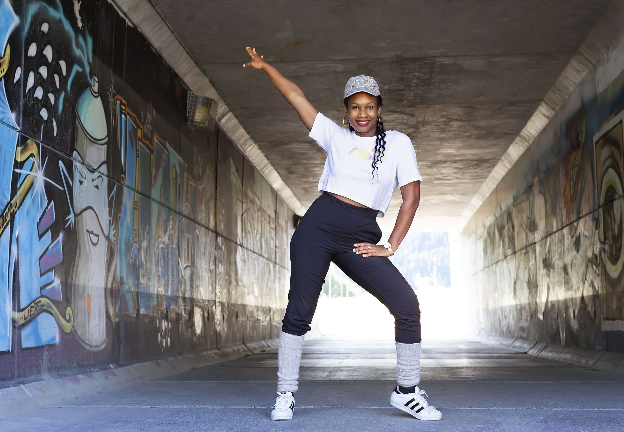 Black woman posing confidently in a graffiti-covered underpass, wearing a gray baseball cap, white cropped t-shirt, black jogger pants, and white sneakers with black stripes, her right hand on her hip and her left arm raised in the air in Norway.