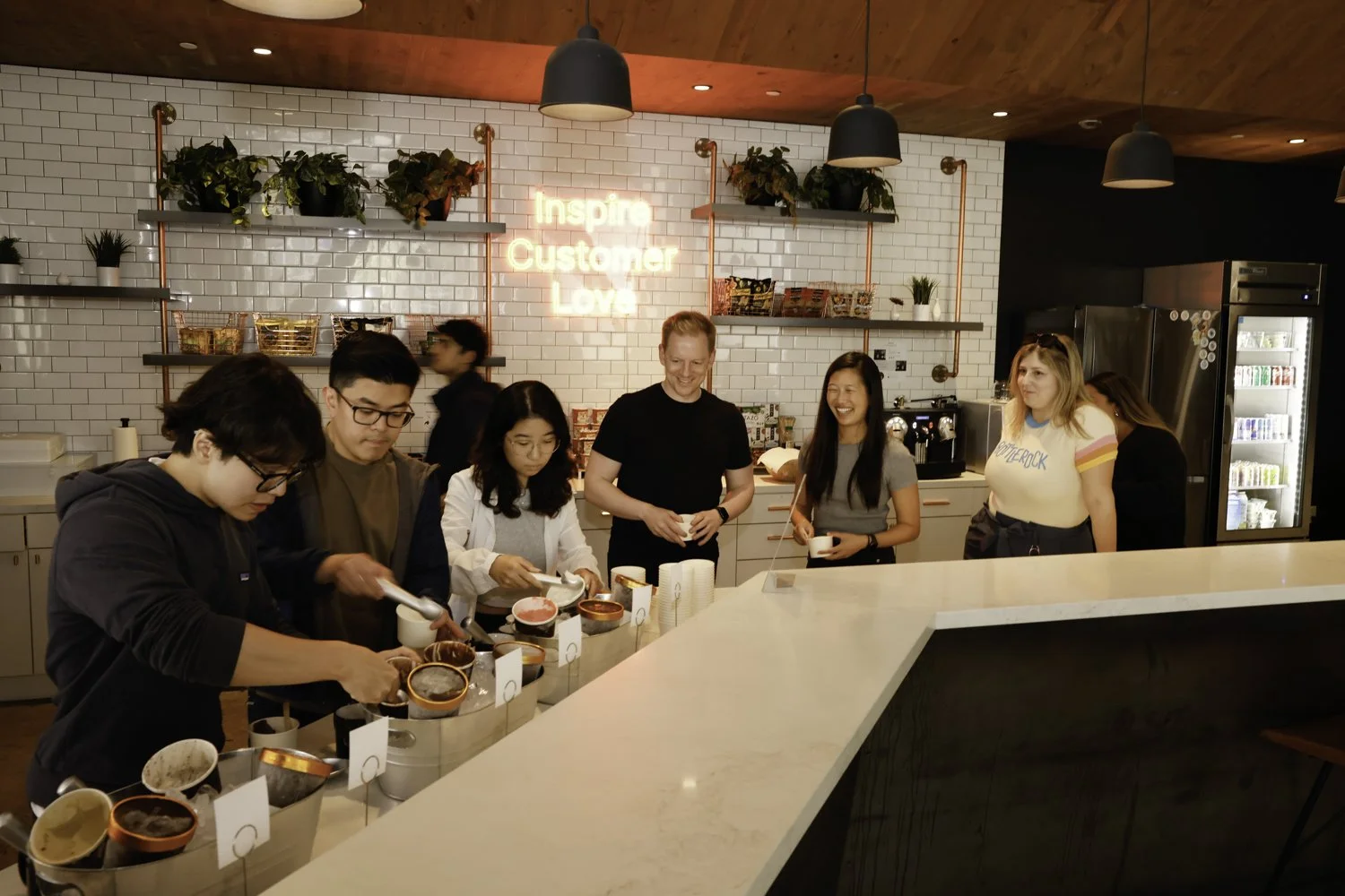 People in line at a coffee shop counter, serving themselves from bowls of ice cream, with a neon sign that reads 'Inspire Customer Love' in the background Brex headquarter in San Francisco, CA.