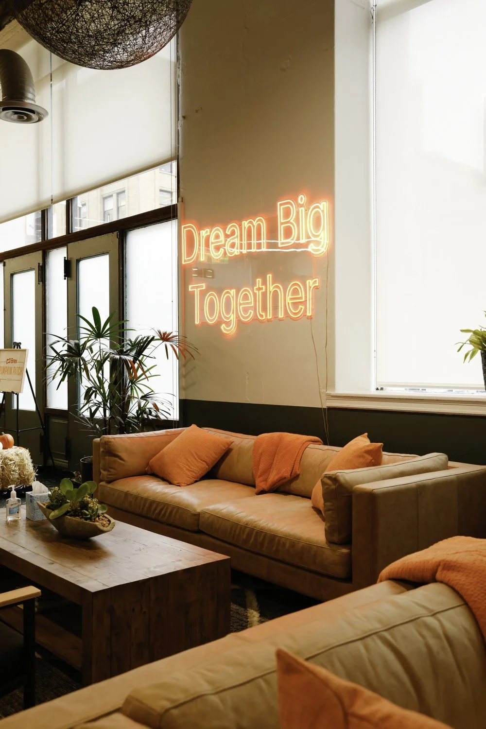 Cozy lounge area with peach couches, orange pillows and blankets, a wooden coffee table with plants, and a neon sign that reads 'Dream Big Together' in a bright indoor space with large windows and plants Brex headquarter in San Francisco, CA.