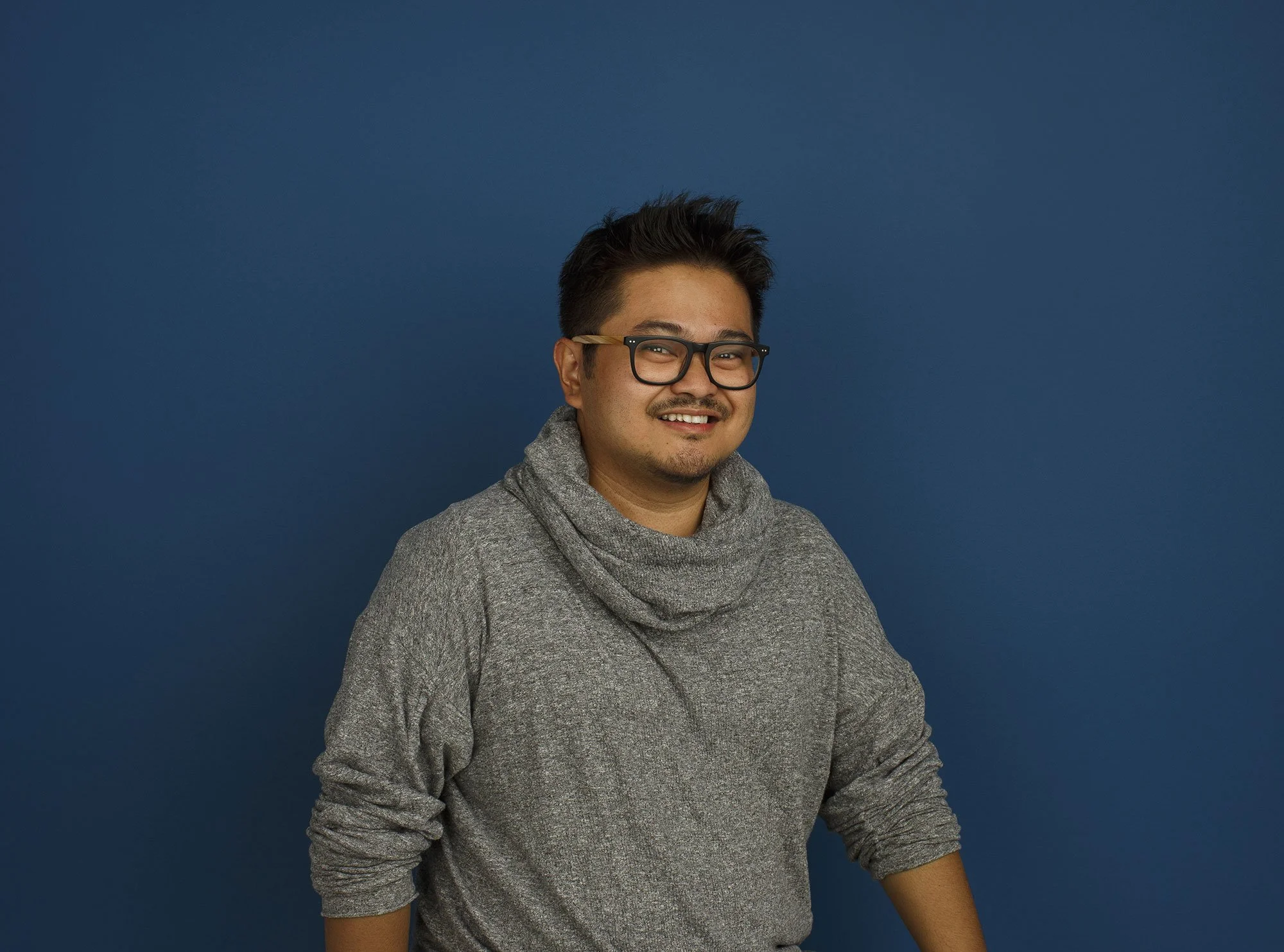 A Phillipino man with dark hair, glasses, and a goatee wearing a gray sweater with a cowl neck, standing in front of a solid blue background, smiling in an architectural office in San Francisco, CA.