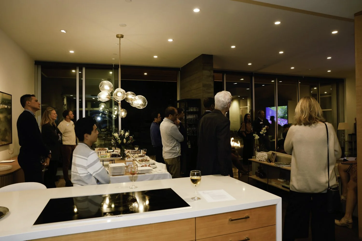 People attending a social gathering or party in a modern living room with a kitchen island, featuring a table with food, a wine glass, and a floral centerpiece. There are large windows, a lighting fixture, and a fireplace, with people standing and ch