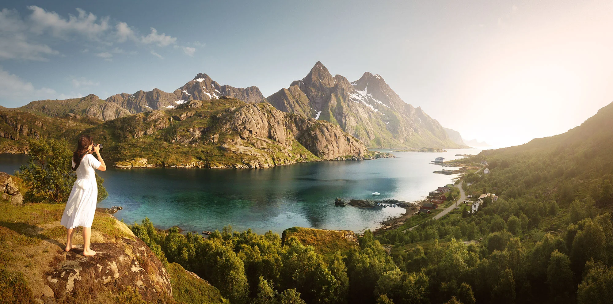 A white woman in a white vintage dress with long brown hair standing on a large rock taking a photo of a scenic landscape with mountains, a lake, green trees, and a small village in Lofoten, Norway.