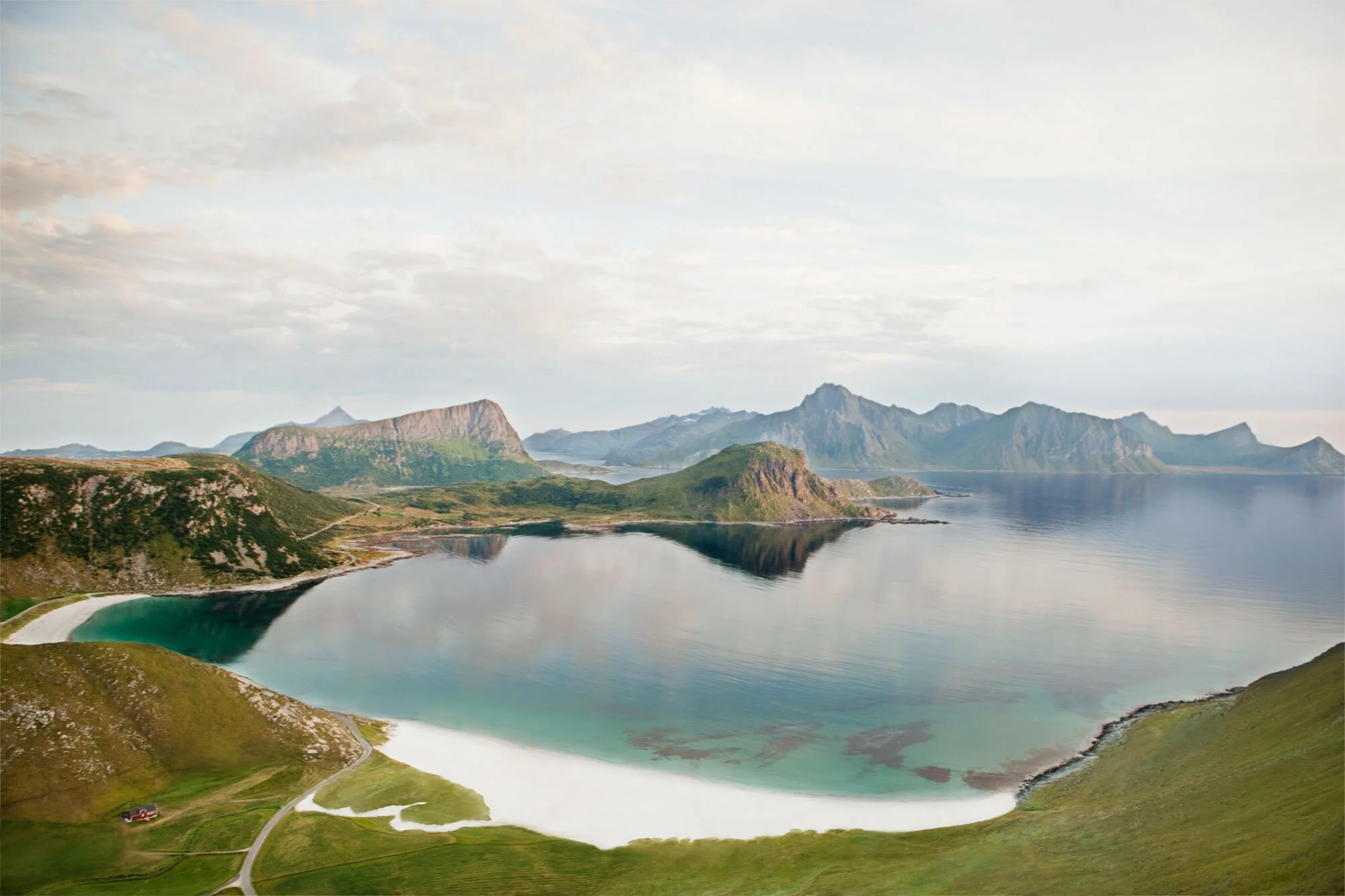 Scenic, panoramic view of a calm, reflective lake surrounded by green hills, a beach, and towering mountains in the background under a cloudy sky in Lofoten, Norway.