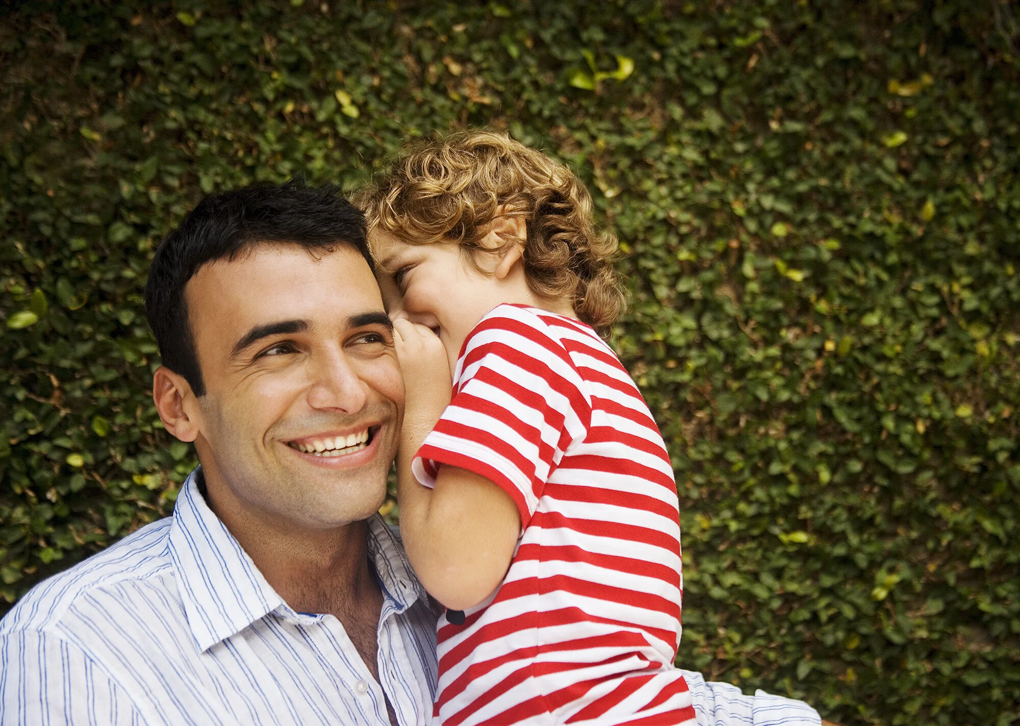 A father smiling with a young child with curly blond hair and striped red and white shirt leaning over him, whispering in his ear in Rio de Janeiro, Brazil.
