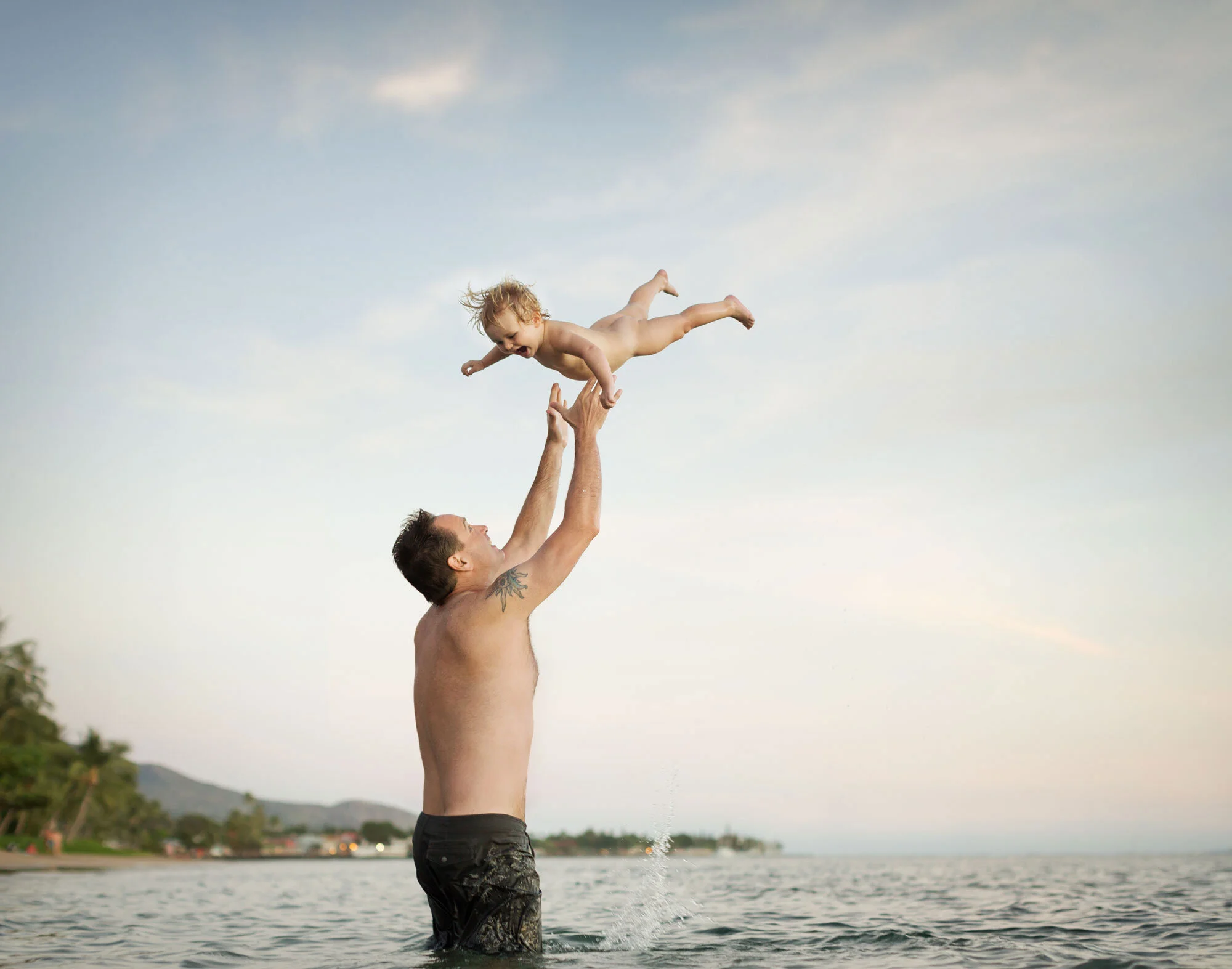 A man is splashing in water at the beach, lifting a toddler into the air. The toddler is smiling and reaching out while the man is looking up at the child, with a scenic background of trees, shoreline, and sky.