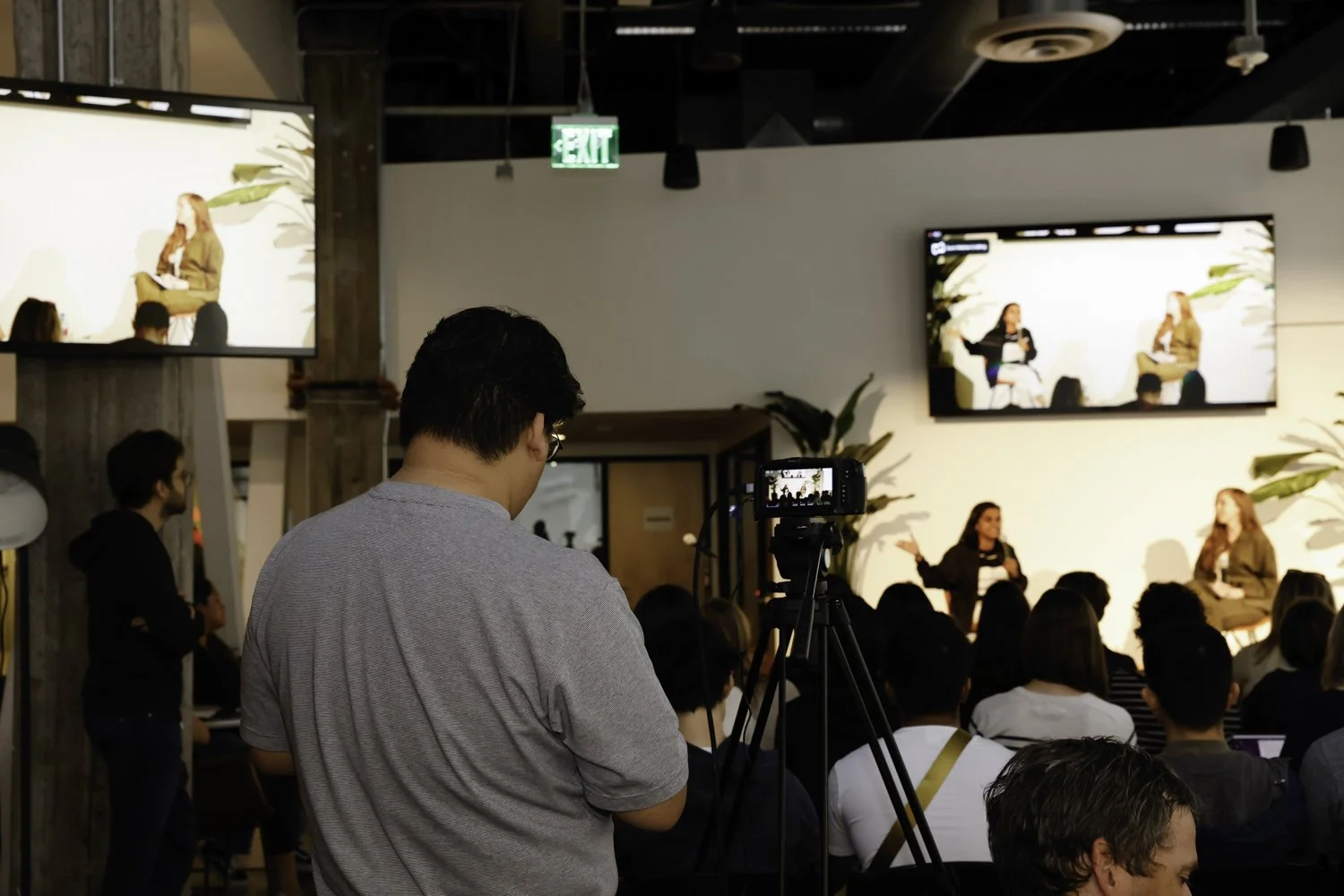 A man films a panel discussion on stage with two women speaking, in a room with an audience, large screens displaying the event, and green exit sign overhead Brex headquarter in San Francisco, CA.