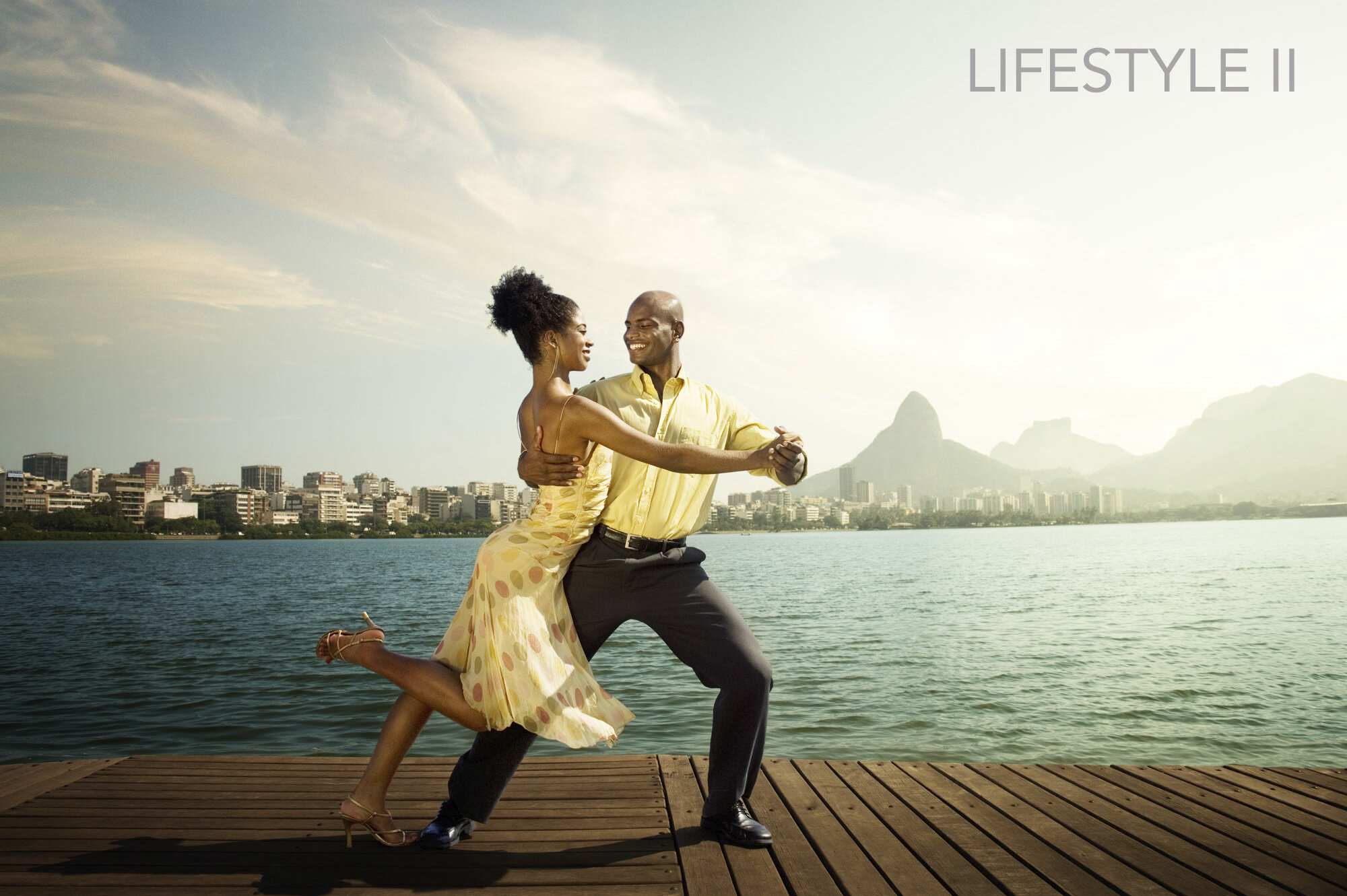 Couple dancing on a wooden pier by the water with a city skyline and mountains in the background, during sunset. The woman is wearing a yellow dress with polka dots, and the man is dressed in a yellow shirt and dark pants at Lagoa in Rio de Janeiro.