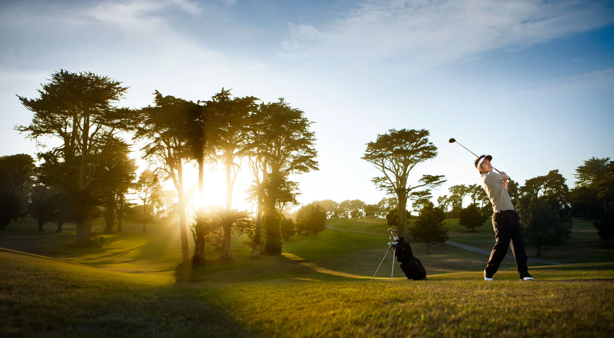 An Asian man playing golf on a course during sunset, with Tee and golf bag in view, surrounded by trees and open sky in the Presidio, San Francisco, CA.