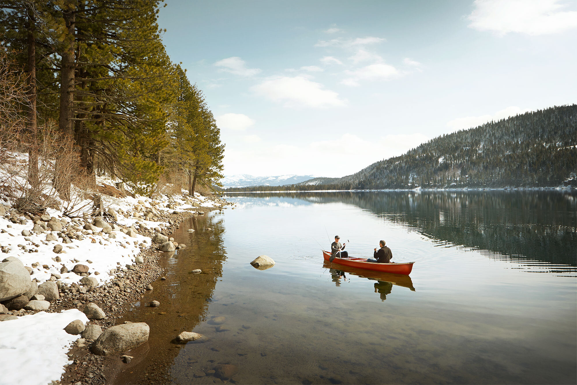 Two people in a red canoe fishing on a calm lake with rocky shoreline, snow, evergreen trees, and forested hills in the background at Donner Lake, Lake Tahoe, CA.