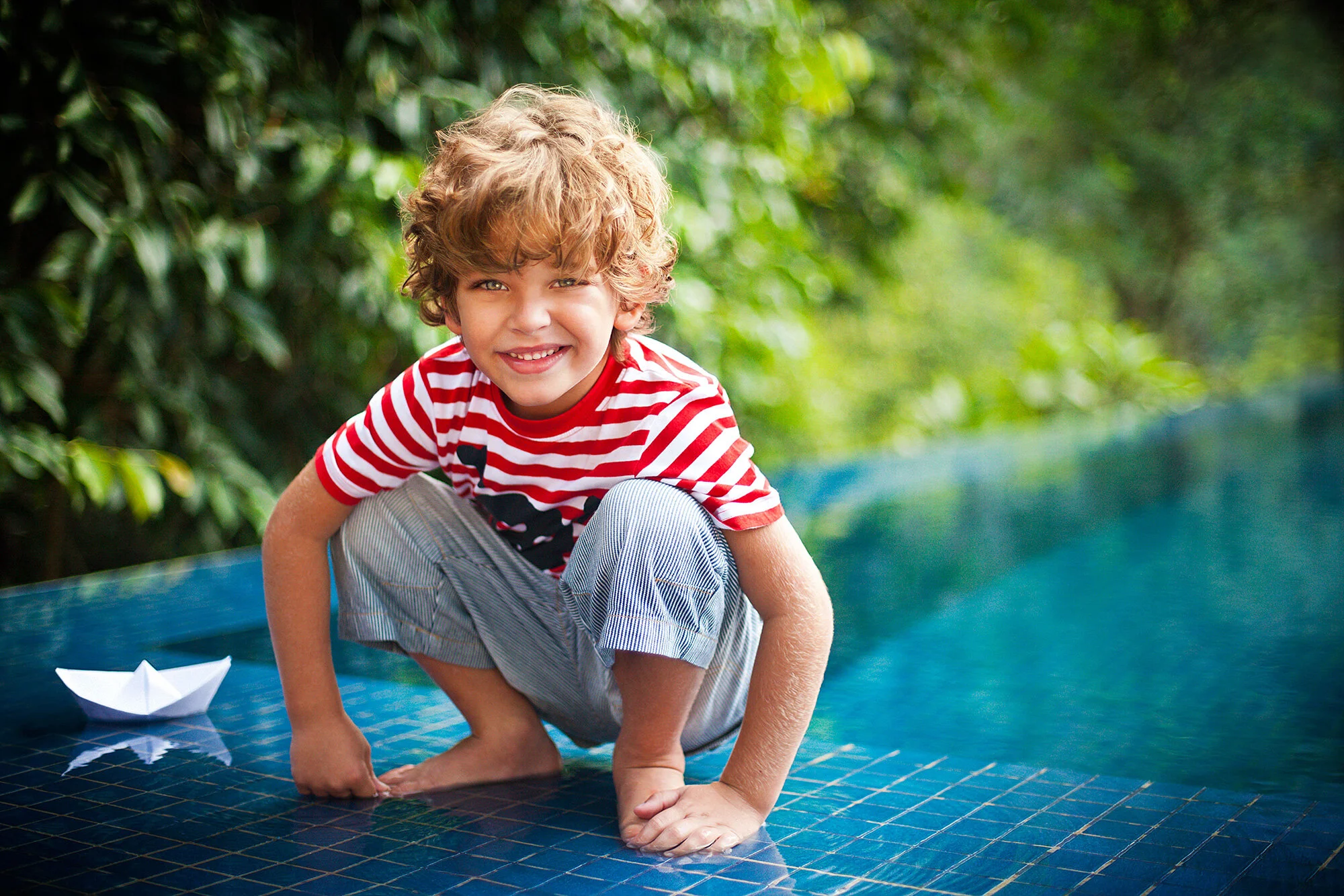 Young Brazilian boy with curly blonde hair smiling and crouching on a poolside tile surface, with a paper boat nearby and green foliage in the background in Rio de Janeiro, Brazil.