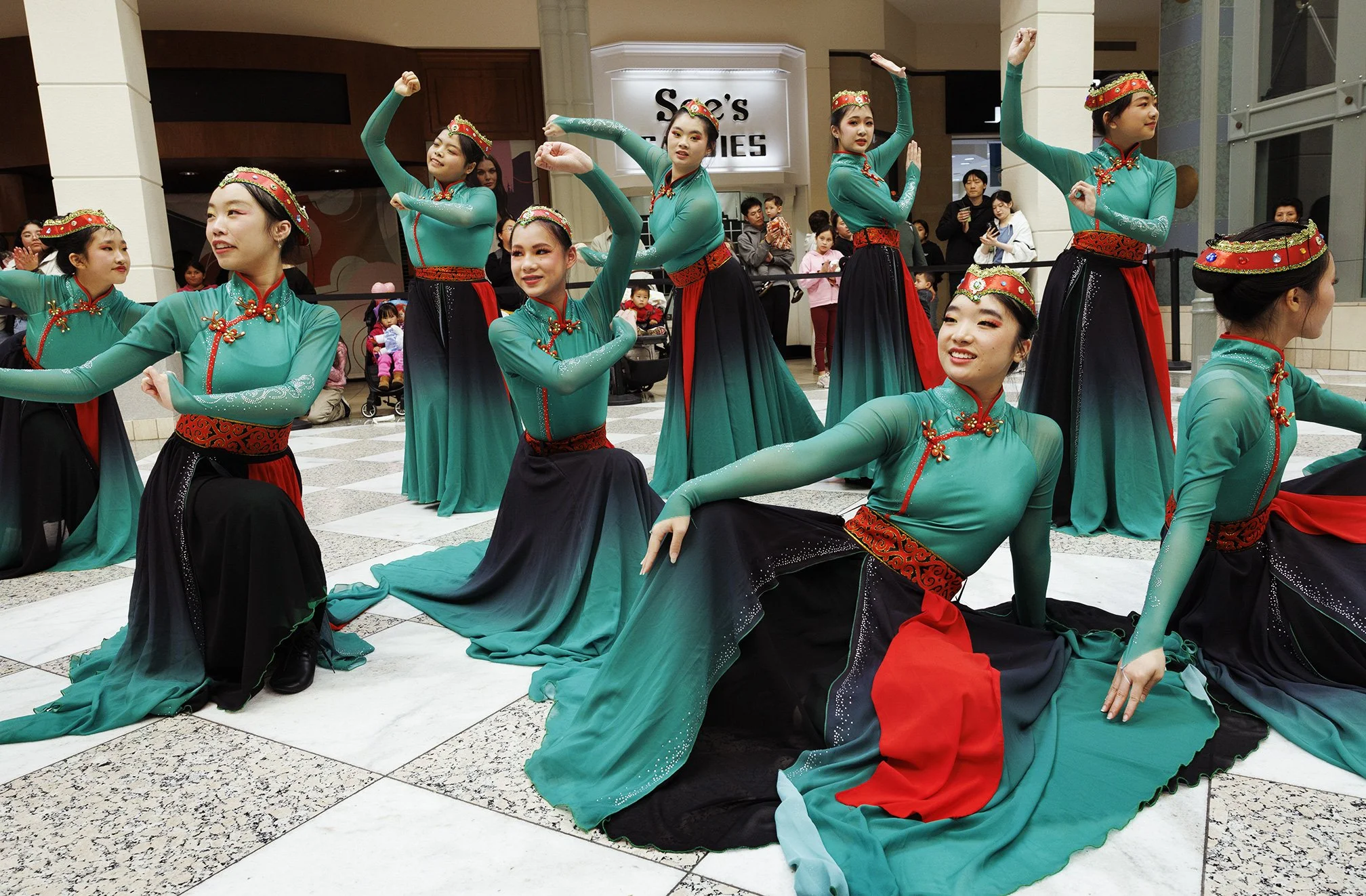 Group of nine Asian women in teal and black traditional dresses performing a dance at a mall in San Francisco for the Lunar year celebrations. They are in various poses, some kneeling, some standing, surrounded by spectators.
