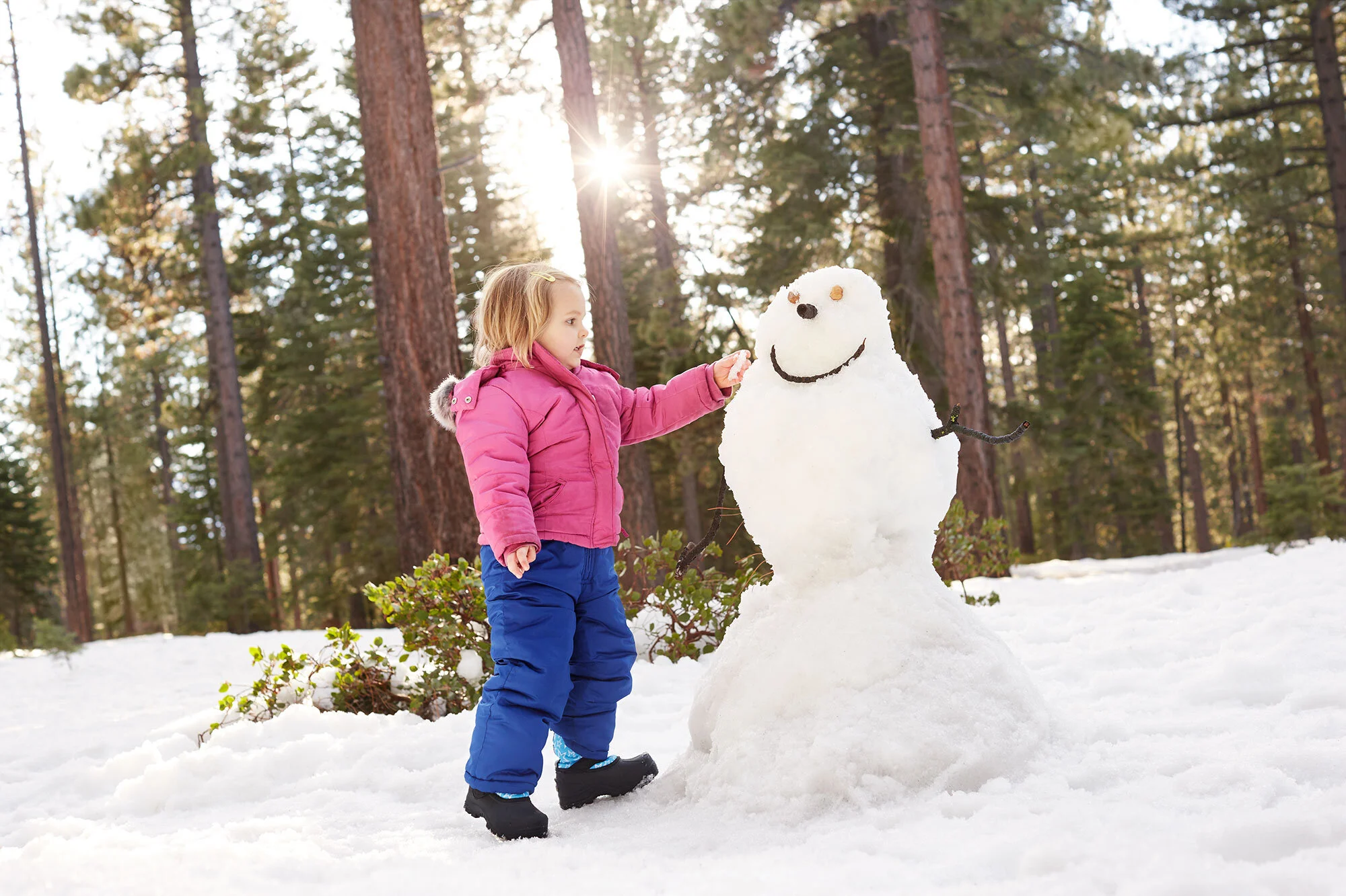A young girl in a pink winter coat and blue snow pants building a snowman in a snowy forest with tall trees and sunlight at Lake Tahoe, CA.