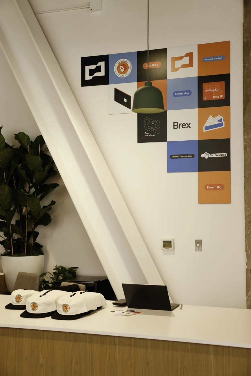 Reception desk with branded hats, laptops, and promotional materials, behind a white counter. Large wall mural with geometric shapes, text, and logos related to Brex and San Francisco, and a green hanging lamp Brex headquarter in San Francisco, CA.
