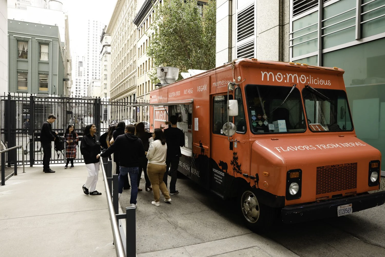 A bright red food truck named "momo:licious" is parked on a city street with several people standing in line to order. The truck has text on it indicating it offers Nepalese flavors, is gluten-free, vegan, and contains no MSG. There are tall building
