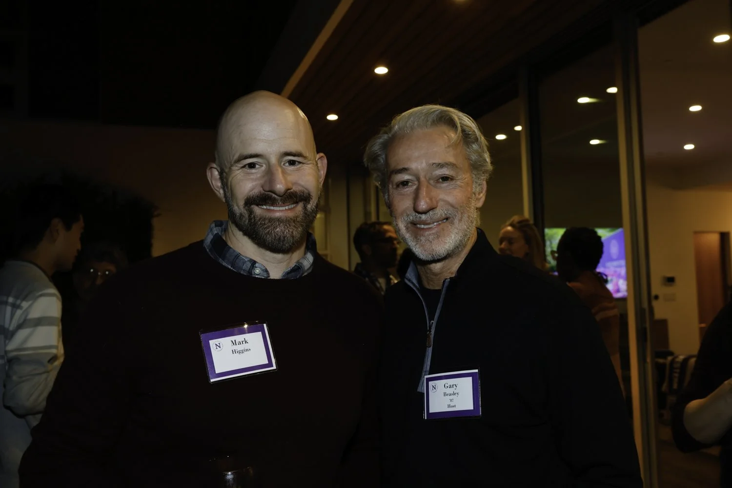 Two men with name tags smiling at an indoor social event. The man on the left has a bald head, beard, and is wearing a dark sweater with a checked shirt underneath. The man on the right has gray hair, beard, and is wearing a black zip-up jacket. Back