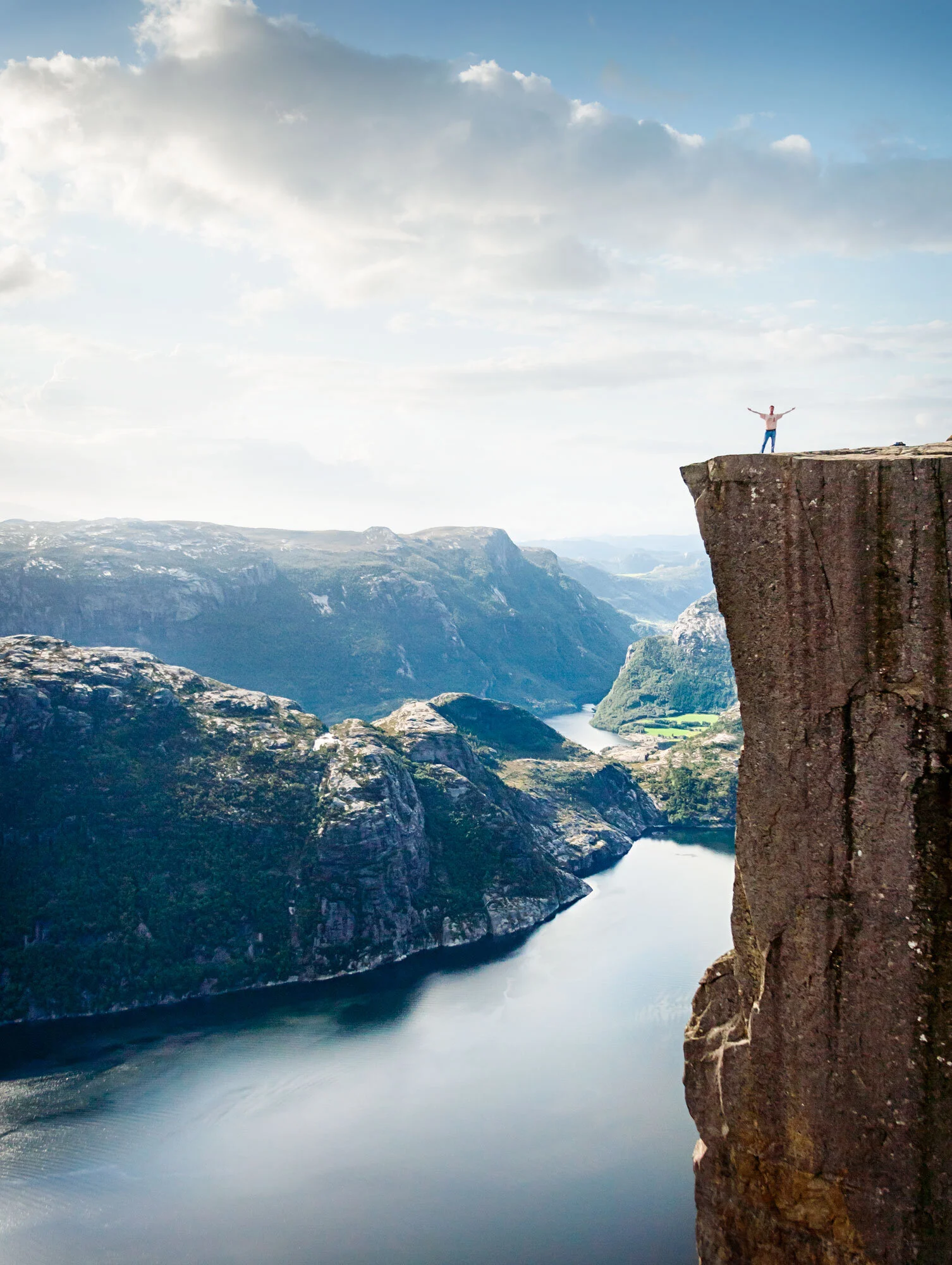 Man standing on top of Pulpit Chair with a fjord underneath about 1000 meters down in Norway with blue sky and clouds.
