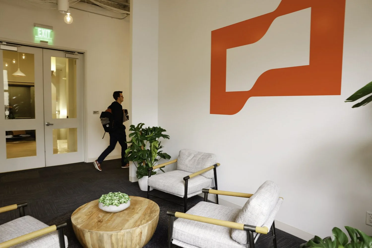 A man walking into a modern office lobby with white walls, a large red logo, and comfortable chairs around a round wooden table with a green plant centerpiece Brex headquarter in San Francisco, CA.