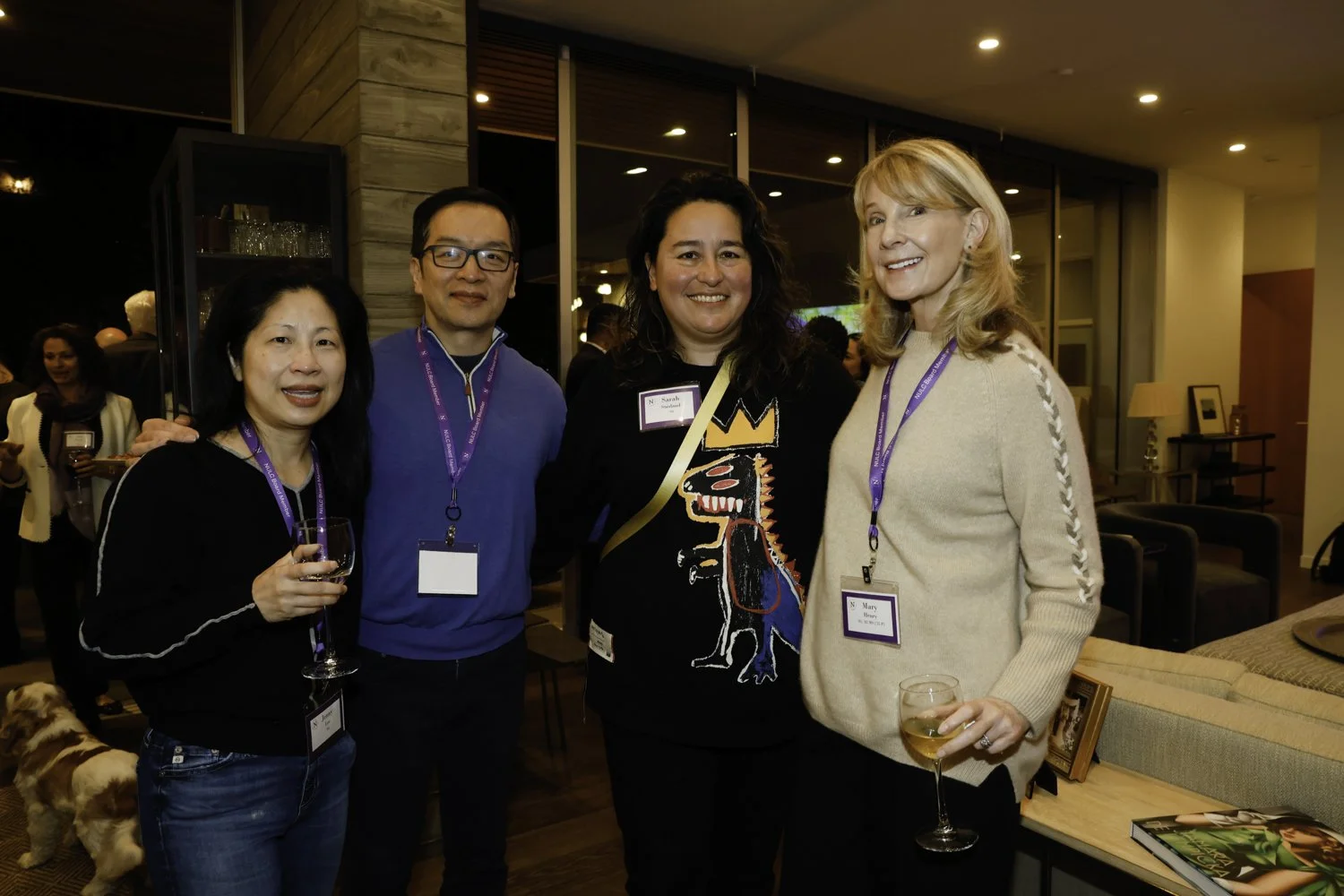 Four people standing together at a social event, smiling at the camera, with two women holding glasses of wine, in an indoor setting with warm lighting, a wooden wall, and other guests in the background.