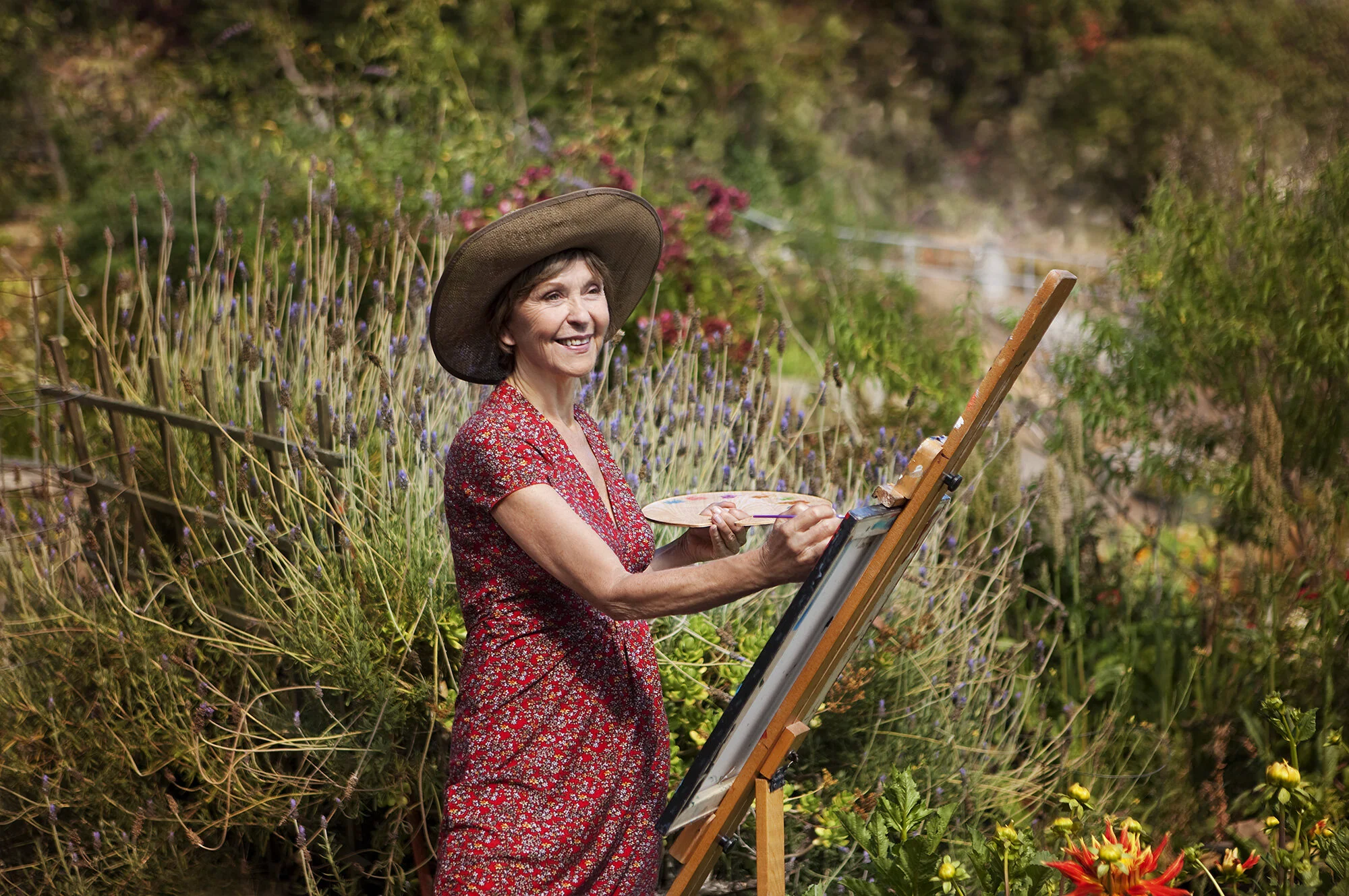 An elderly white woman in a red floral dress and large sun hat painting on a canvas outdoors in a lush community garden in San Francisco, CA