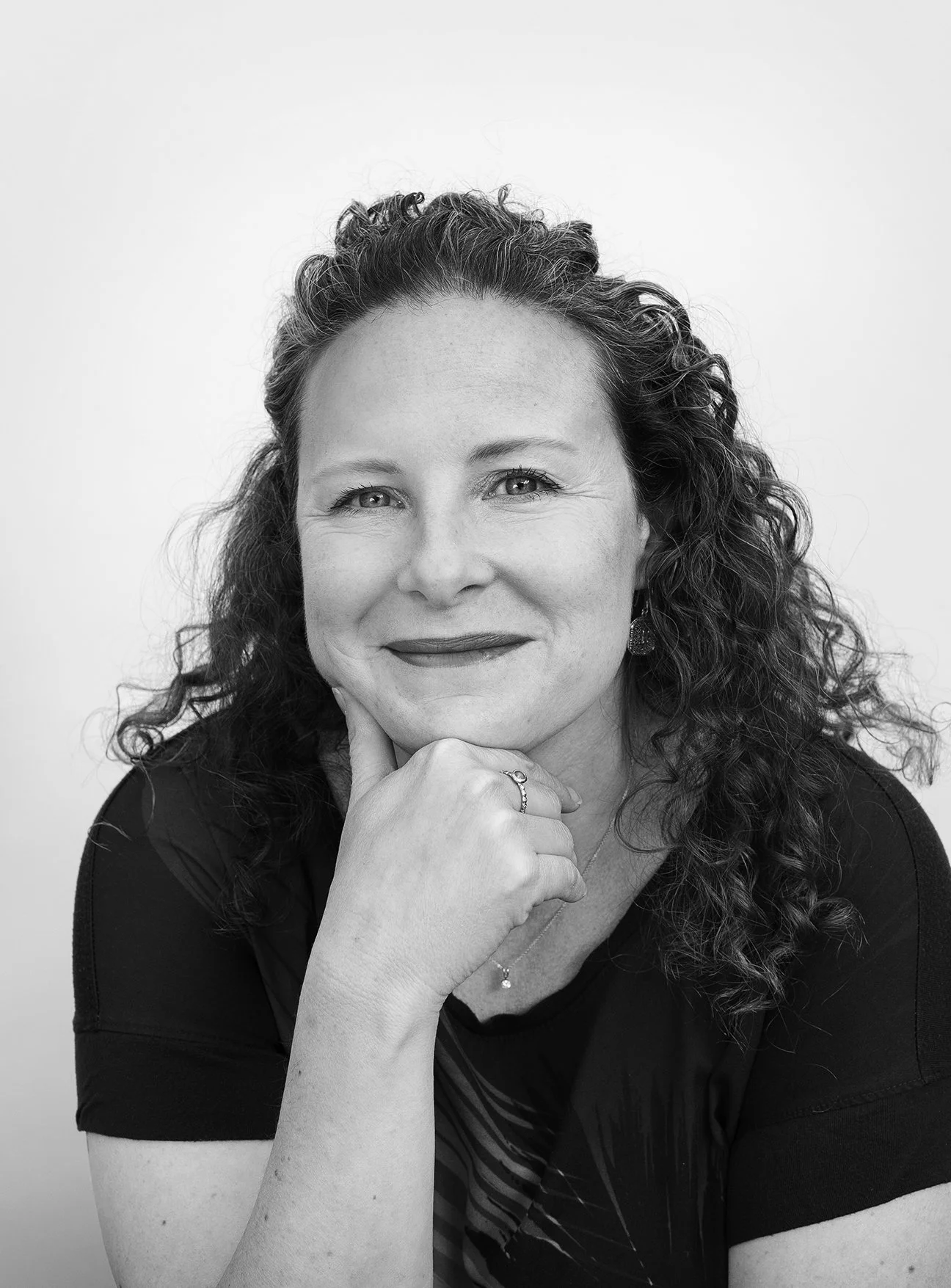 A white woman with curly hair holding her chin with her hand, wearing a black, short sleeved shirt smiling lightly towards the camera in San Francisco, CA.