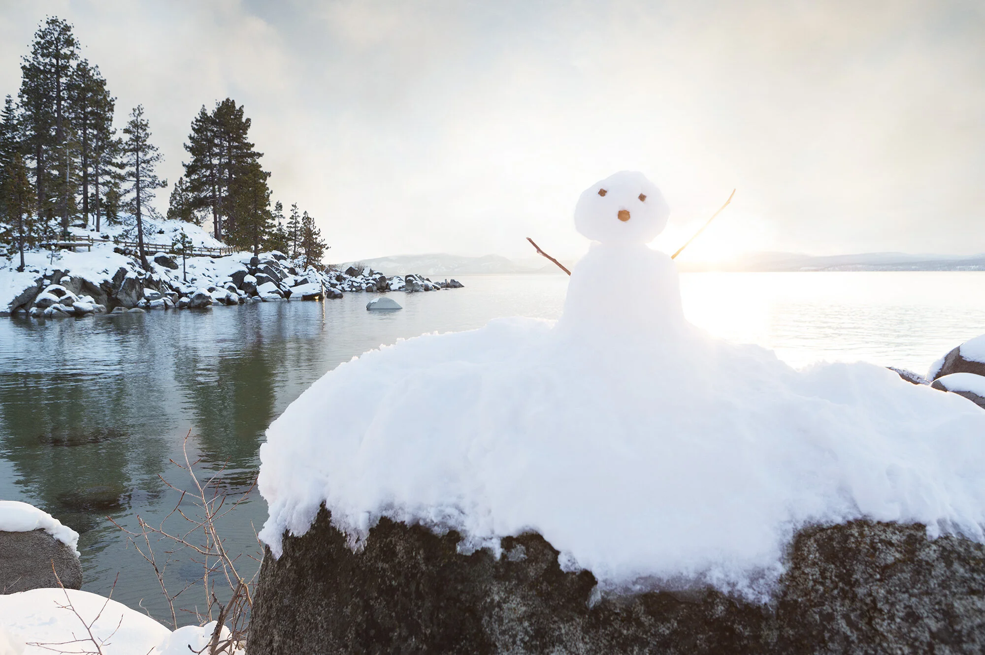 A small snowman on a large rock near a body of water with snow-covered trees on a lakeshore; the sun is setting or rising, creating a bright background at Sand Harbor, Lake Tahoe, CA.