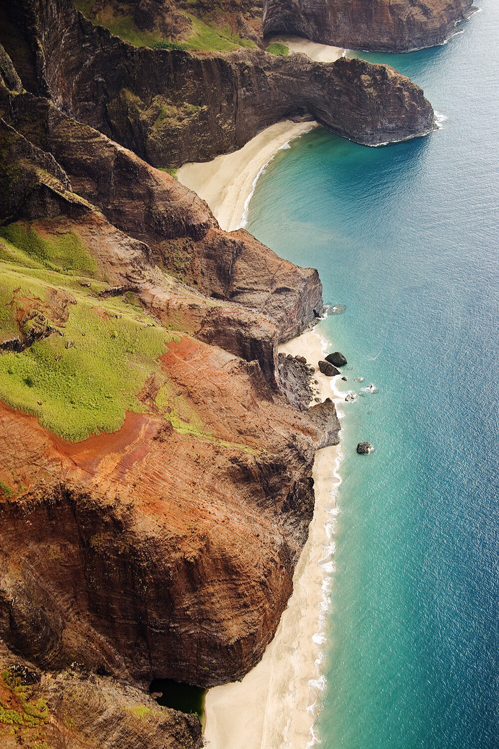 Aerial view of rugged cliffs and green landscape along a sandy beach with turquoise ocean water in Kaua'i, Hawai'i.