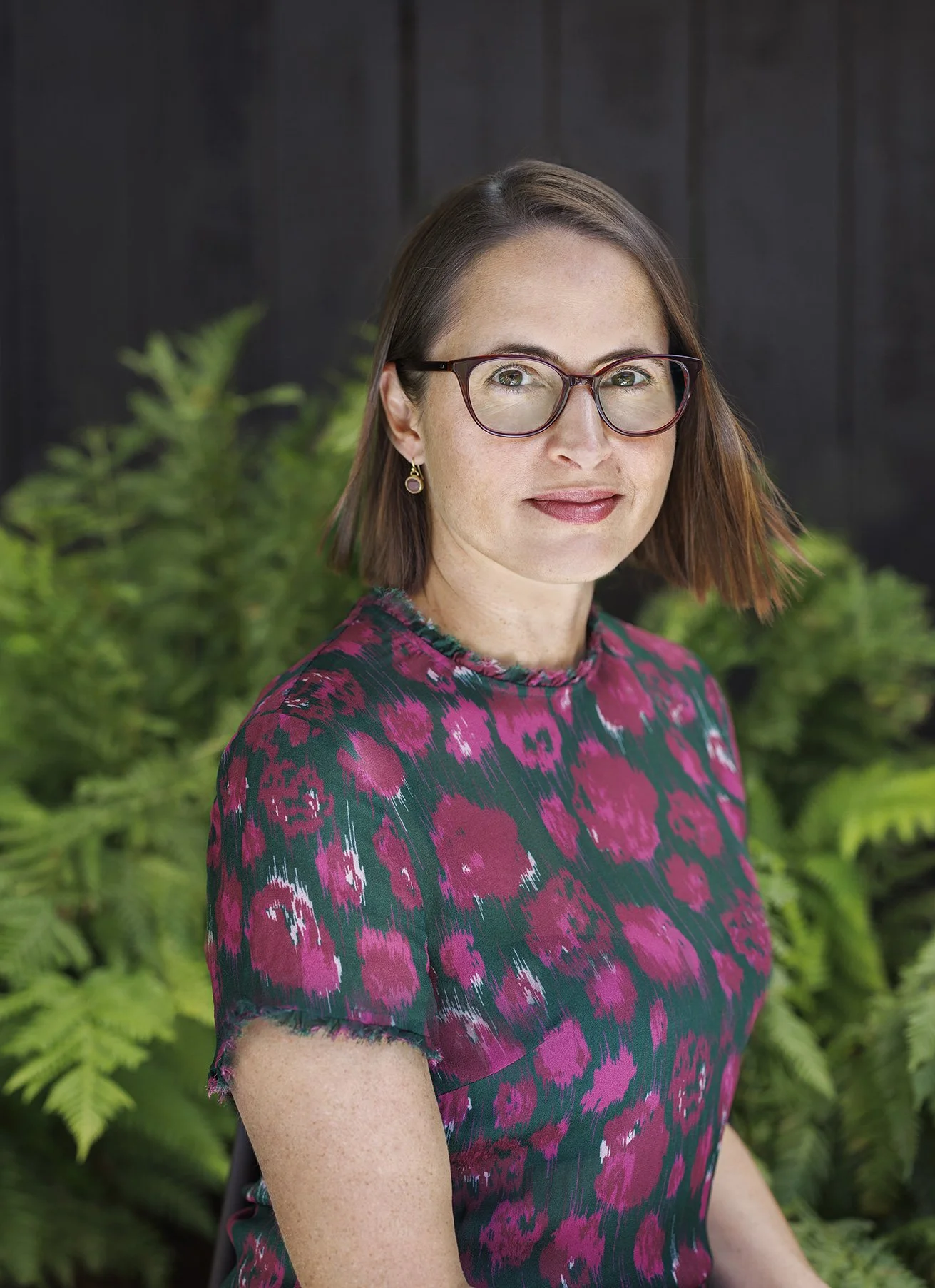 Young Kiwi woman looking at camera at an angle dressed in punk and black shirt, wearing glasses against a green fern and black wooden fence in San Francisco, CA.