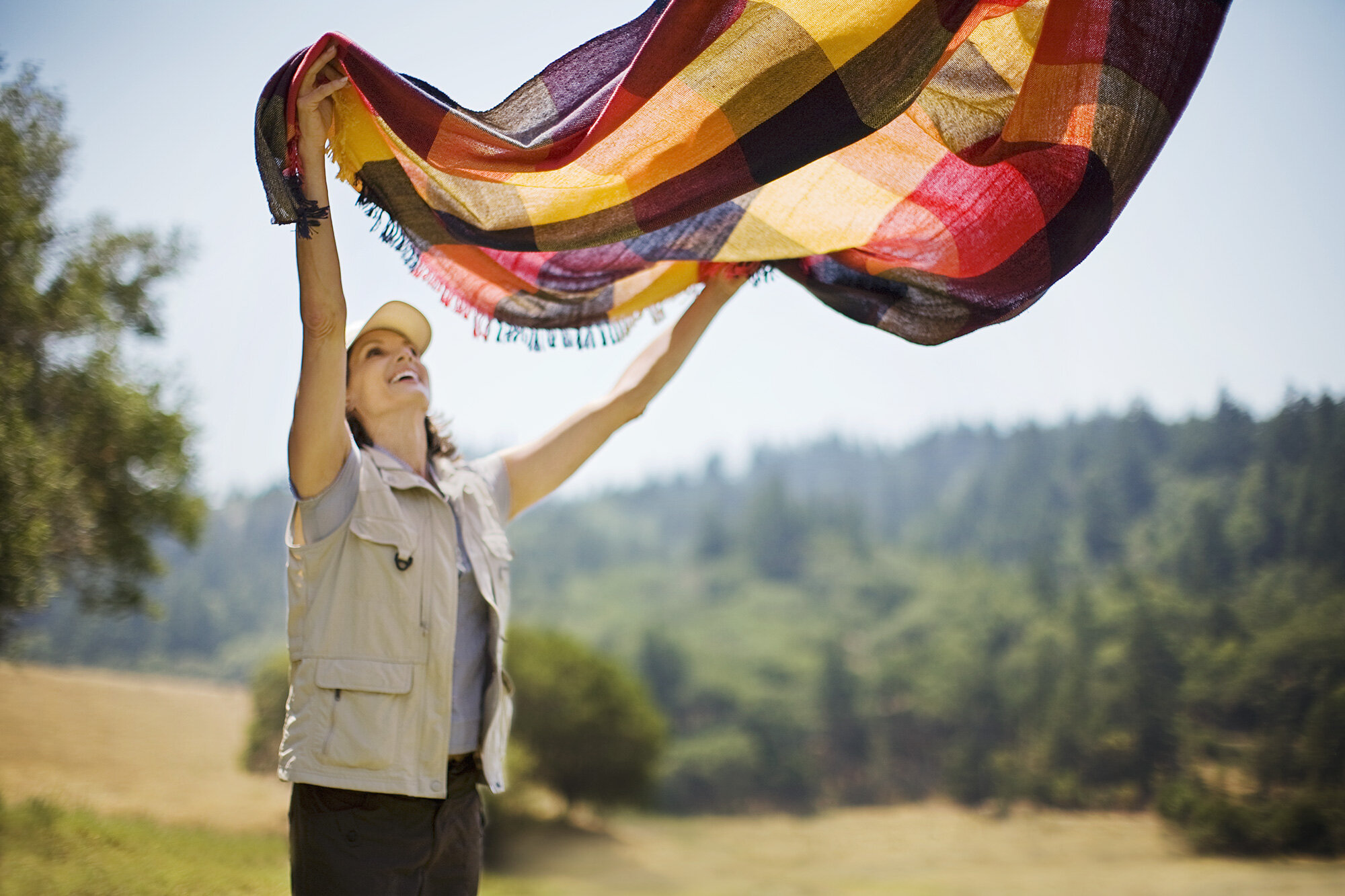 Woman outdoors wearing a beige vest holding a colorful blanket in the air against green trees and a blue sky in Marin, CA.