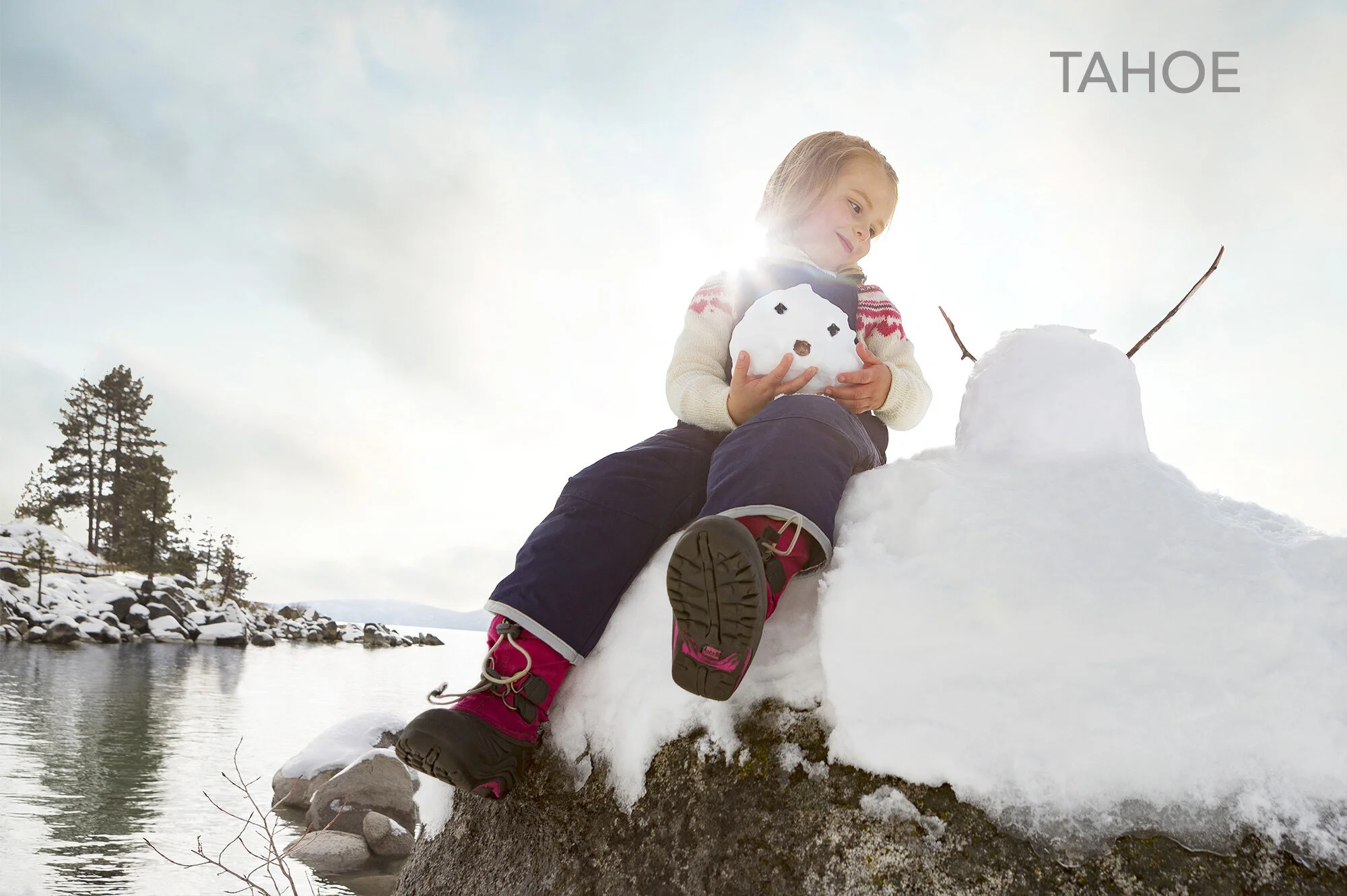 Child sitting on a snowy rock dressed in winter clothing, happily playing with snowman with twig arms near a body of water, holding a small snowhead at Sand Harbor in Tahoe. The background has snow-covered trees and a clear sky with the sun shining. 