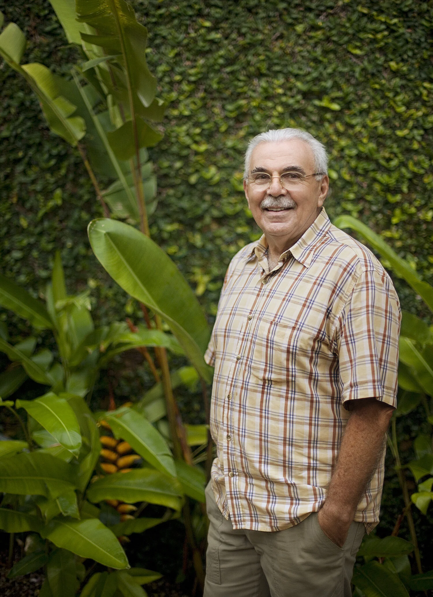 An older Brazilian man with white hair, glasses, and a mustache, smiling in a garden with lush green plants and banana leaves around him. He is wearing a plaid button-up shirt and khaki pants, with his right hand in his pocket in Rio de Janeiro, Braz