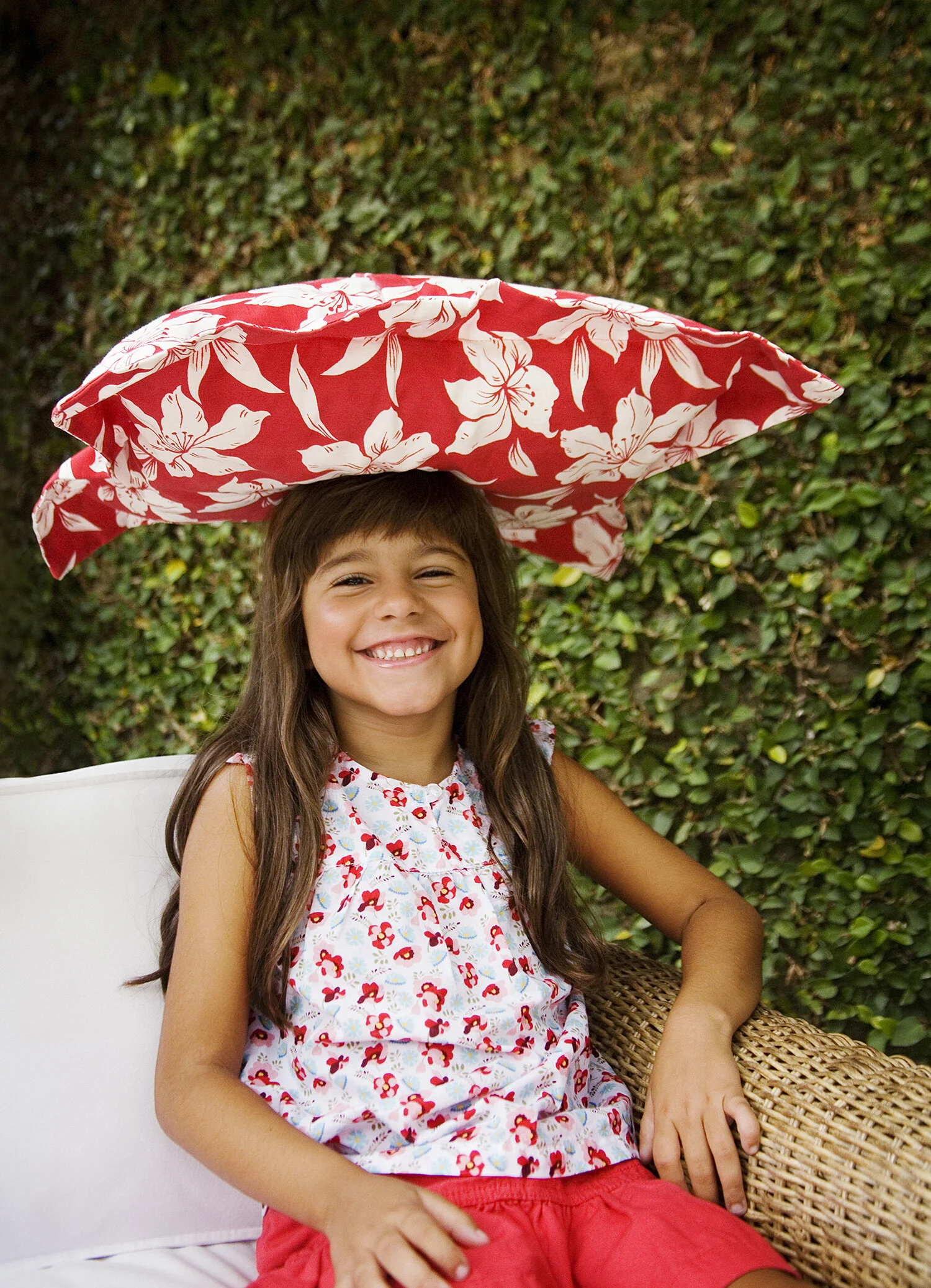 A young girl with long brown hair and a big smile, sitting outdoors under a large red and white floral-patterned umbrella. She is wearing a sleeveless top with a red floral pattern and red shorts, with green foliage in the background in Rio de Janeir